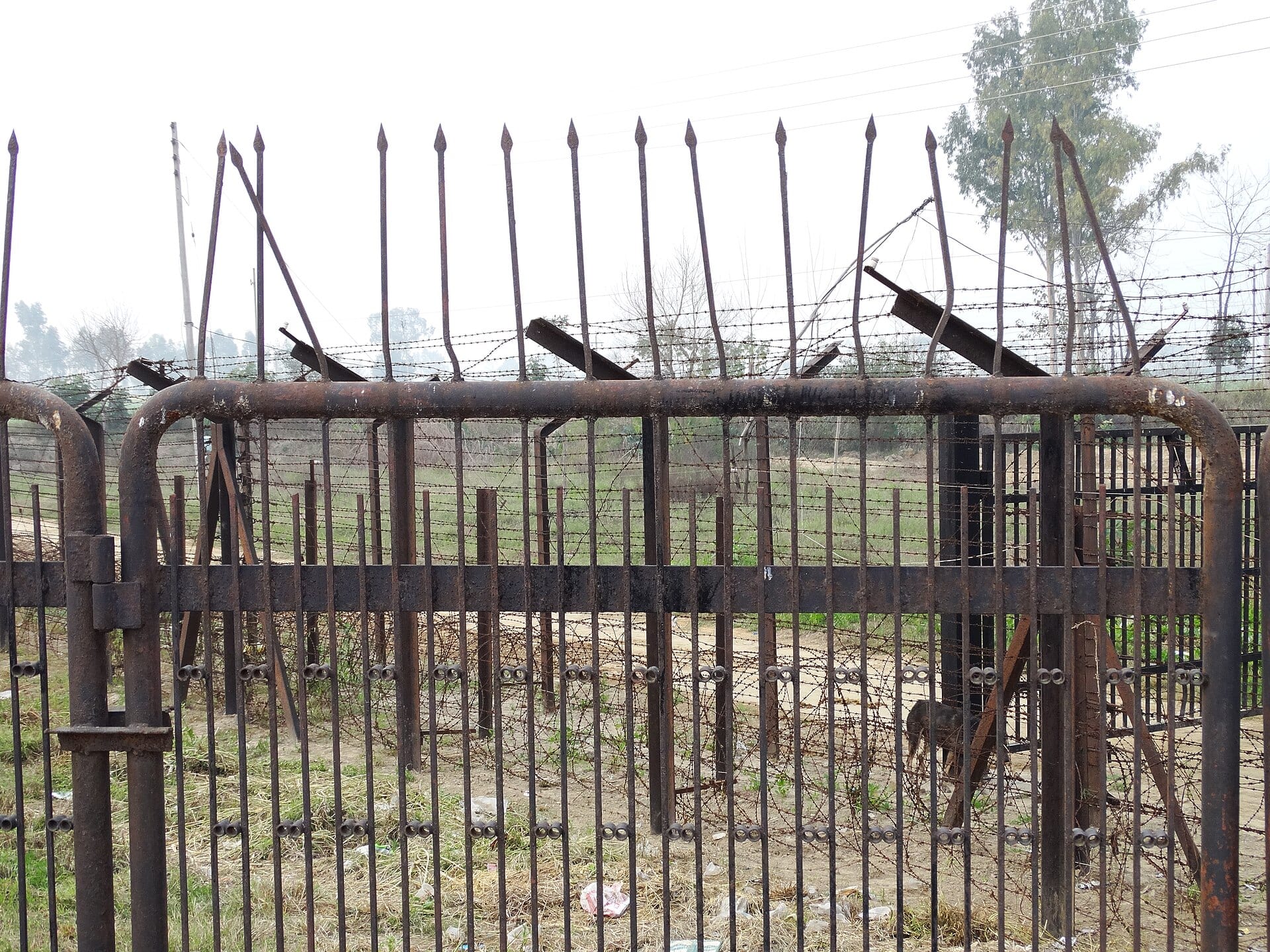 A rusted metal fence with spikes and barbed wire beside a dirt track, with trees behind, at the Attari-Wagah crossing near Amritsar.