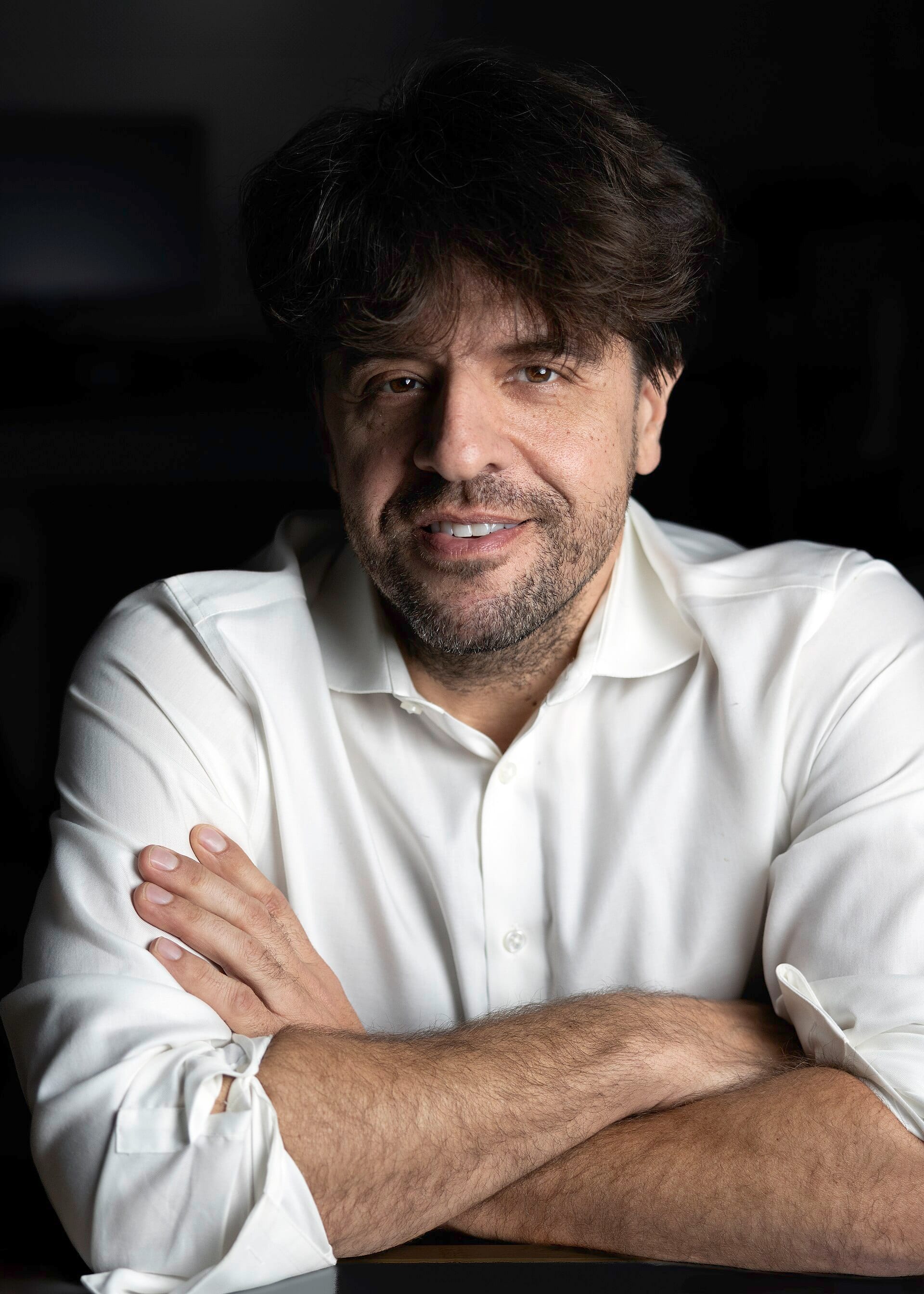 Portrait of a dark-haired man in a white shirt with his arms crossed on a table, against a black background.