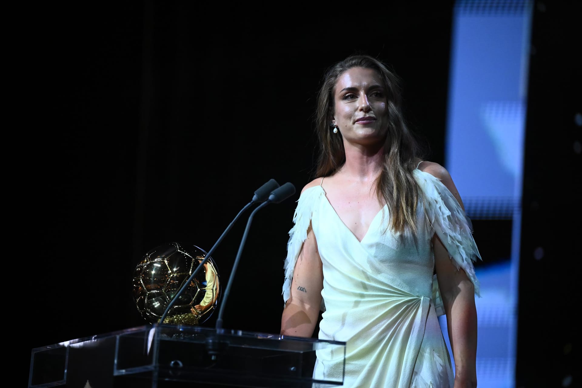 Alexia Putellas, de vestido blanco con mangas de plumas, de pie tras un atril con dos micrófonos y un balón dorado a su lado.