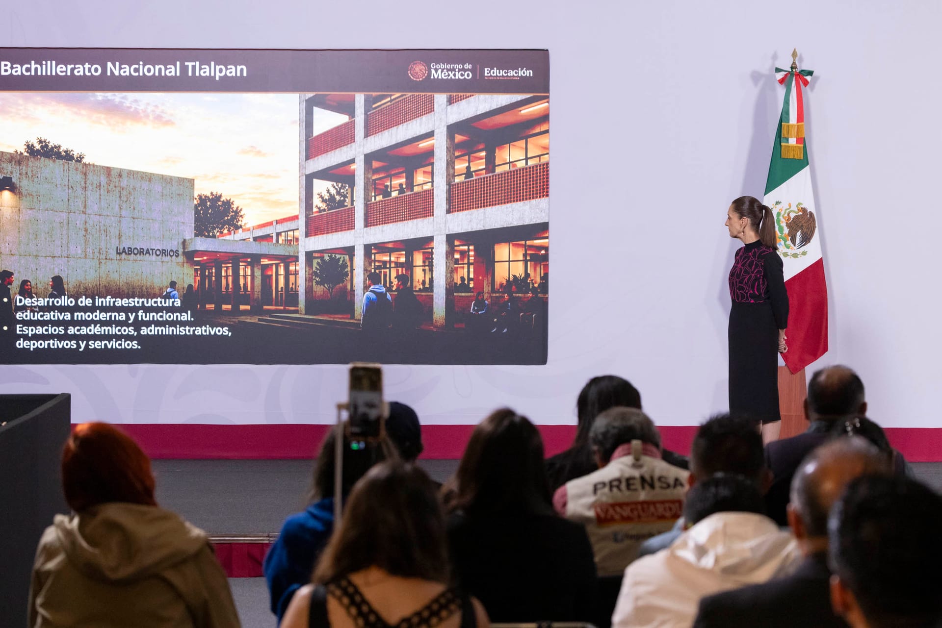 Claudia Sheinbaum, de pie junto a la bandera, mira una lámina del Bachillerato Nacional Tlalpan proyectada ante periodistas sentados.
