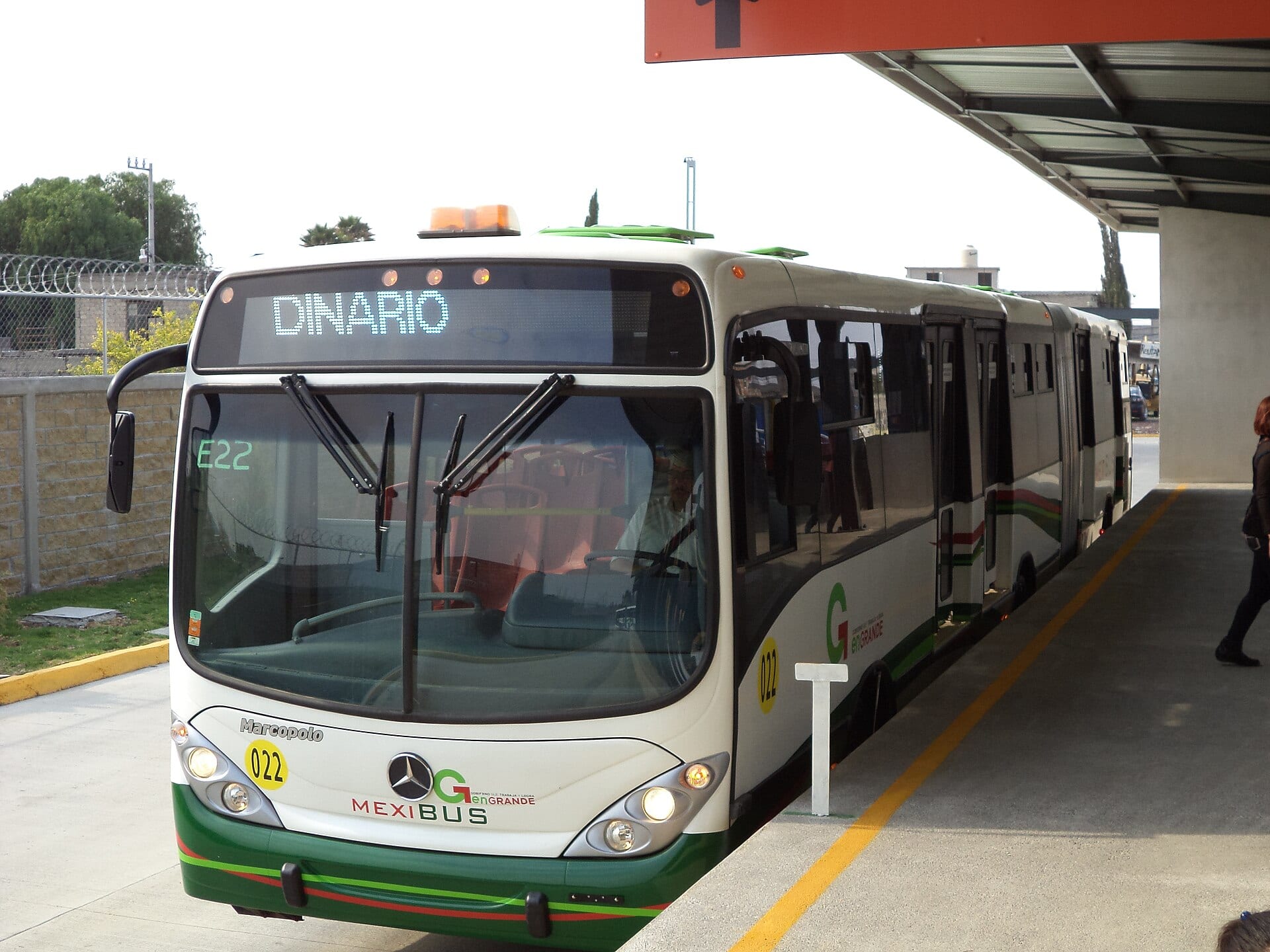 Un autobús articulado blanco y verde del Mexibús, con el letrero Dinario en su pantalla frontal, espera bajo la marquesina de una estación.