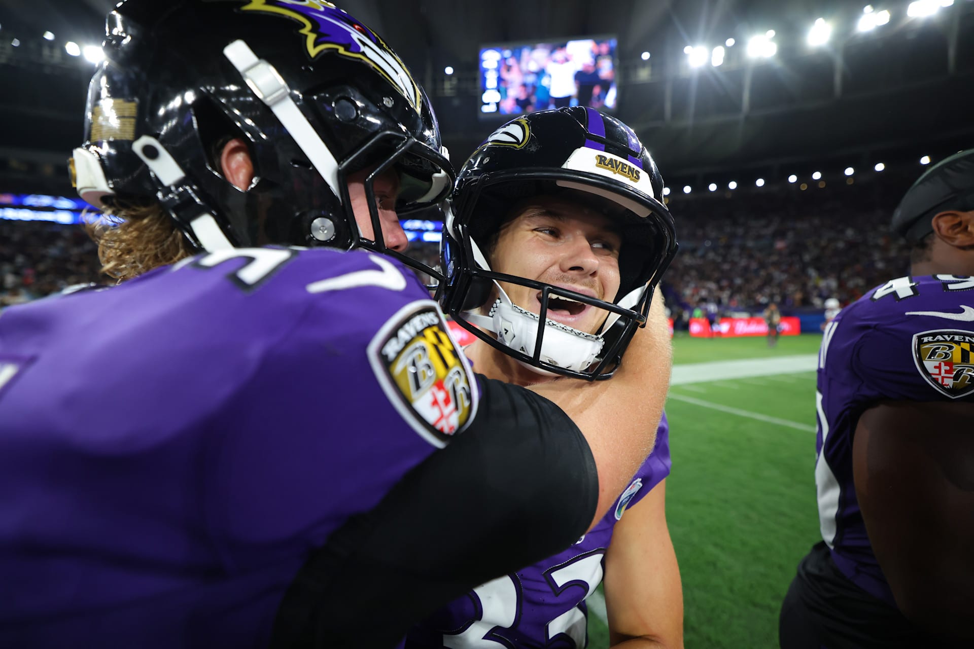 Tyler Loop, pateador de los Ravens con casco negro y camiseta morada, sonríe mientras un compañero lo abraza en el campo del Maracaná.