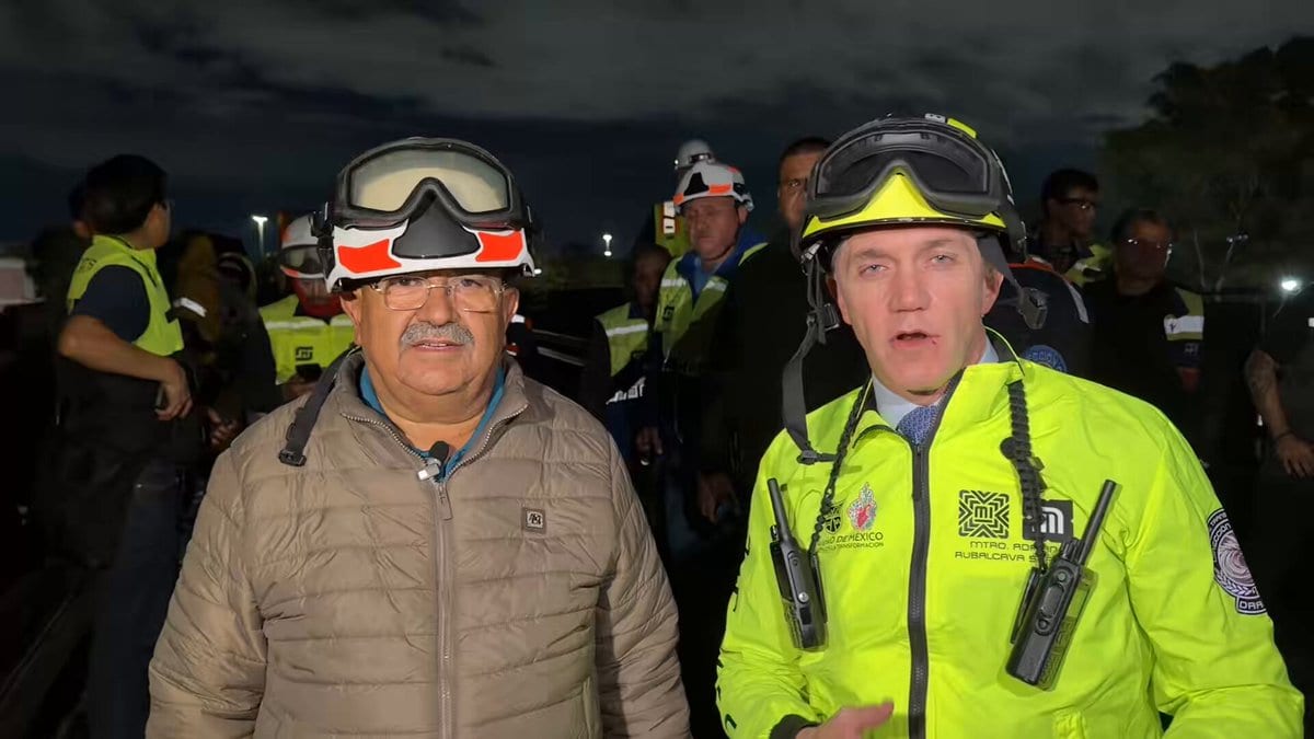 El director del Metro, Adrián Rubalcava, con casco y chaleco amarillo, junto a un hombre con chamarra beige, de noche y rodeados de personal con chalecos reflectantes.