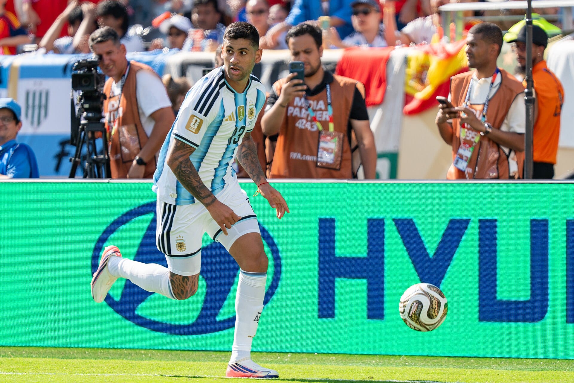 Cristian Romero, con la camiseta albiceleste, corre por la banda con el balón a su derecha ante fotógrafos y aficionados en las gradas.