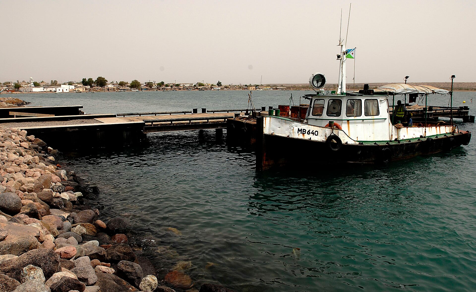 A white and black tugboat moored at a stone pier, with the town of Obock across the water in the background.