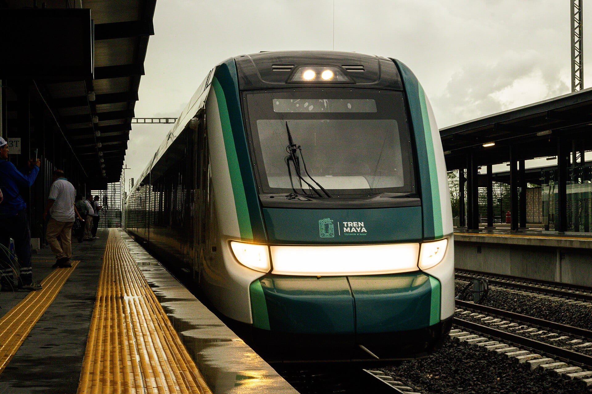 Un tren verde y blanco con el logotipo del Tren Maya llega a un andén mojado mientras varias personas esperan a la izquierda.