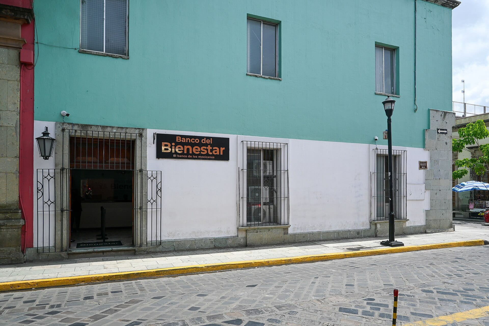 Fachada de una sucursal del Banco del Bienestar en una esquina empedrada, con un letrero negro sobre el muro blanco y la puerta abierta a la izquierda.