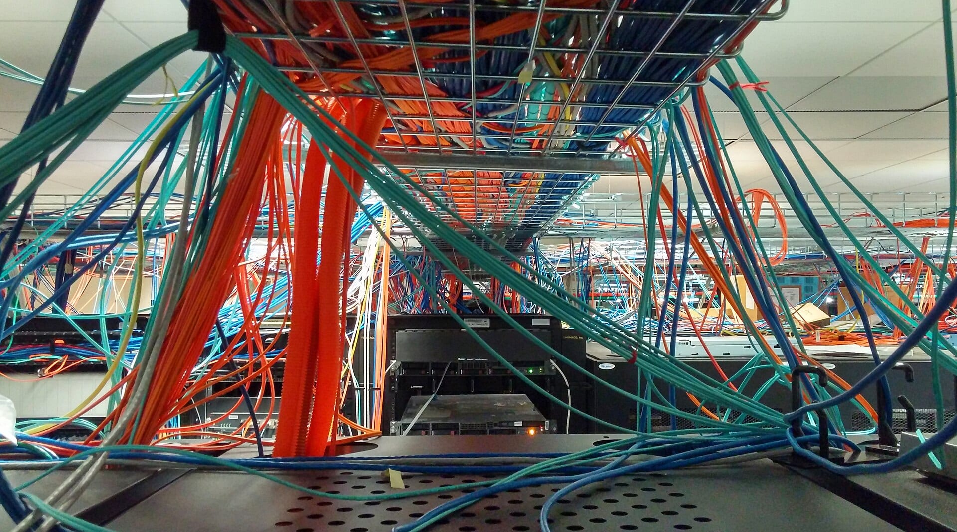 Bundles of blue, green and orange network cables hang from metal trays above equipment in a data center.