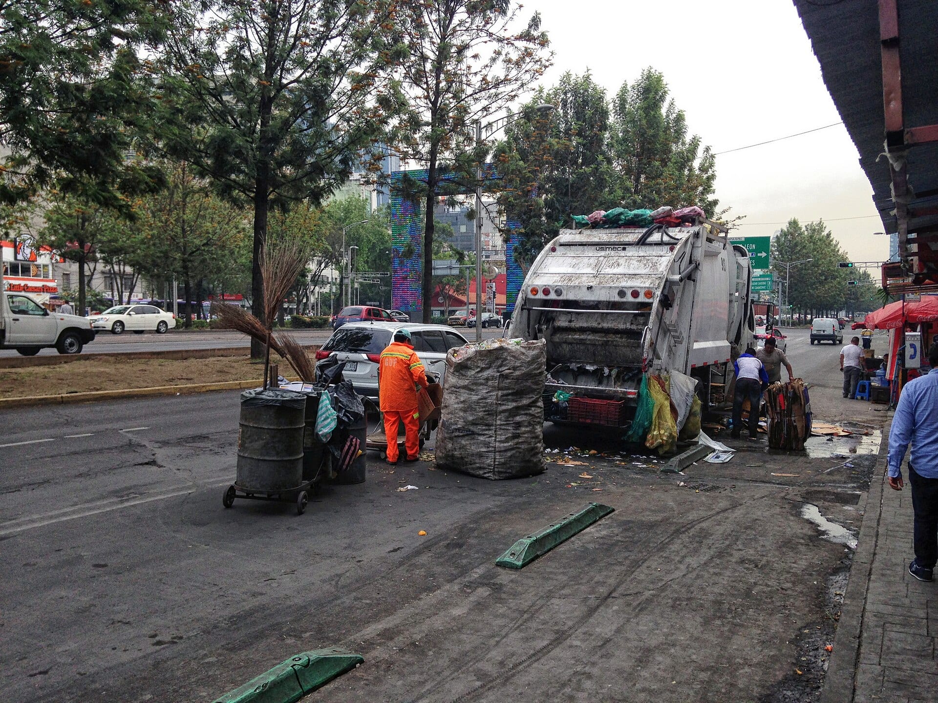 Un camión recolector blanco recibe bultos de residuos en una avenida, junto a un trabajador de uniforme naranja.