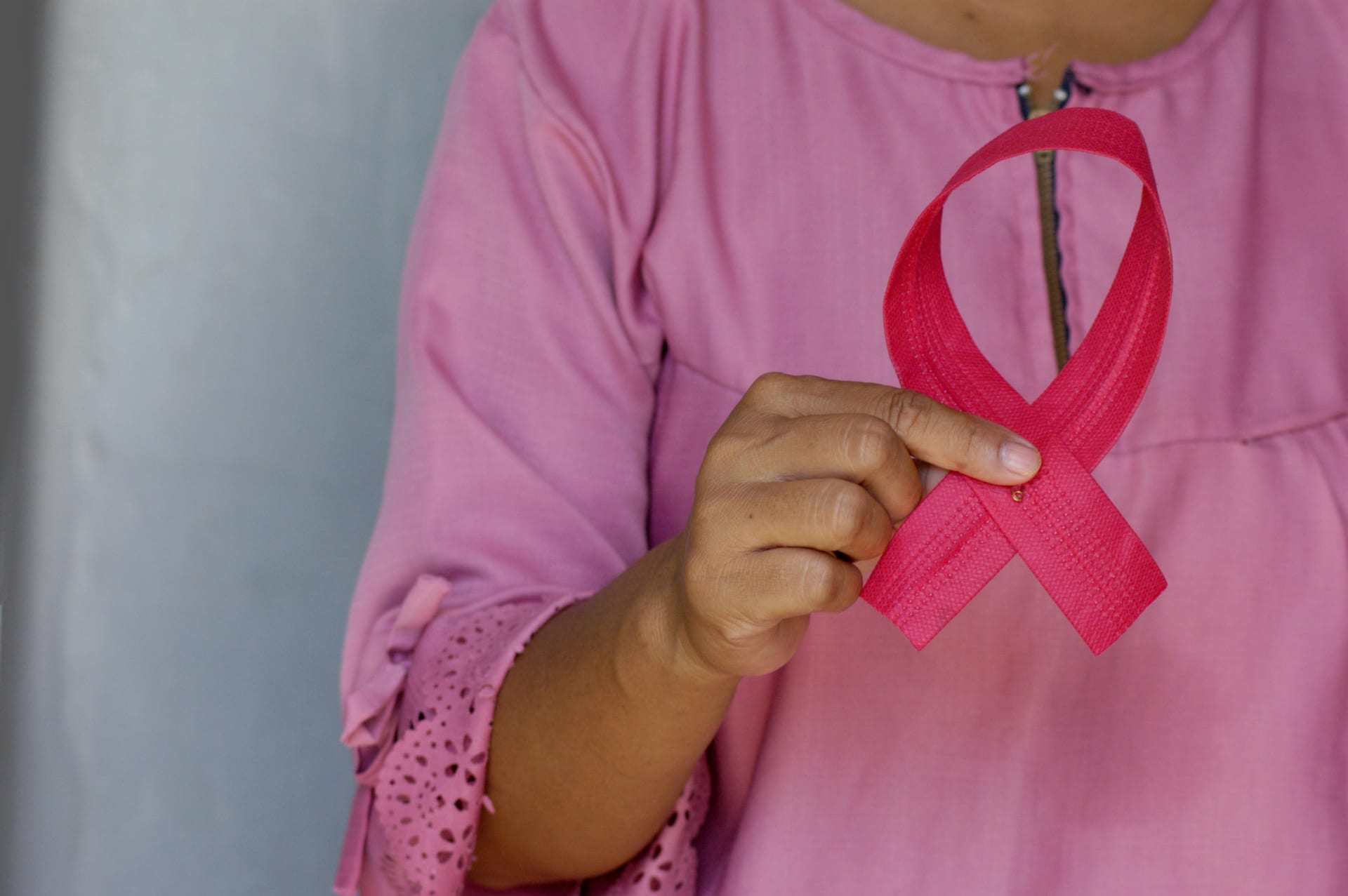 Una mujer con blusa rosa sostiene frente a su pecho un listón rosa doblado en forma de lazo.