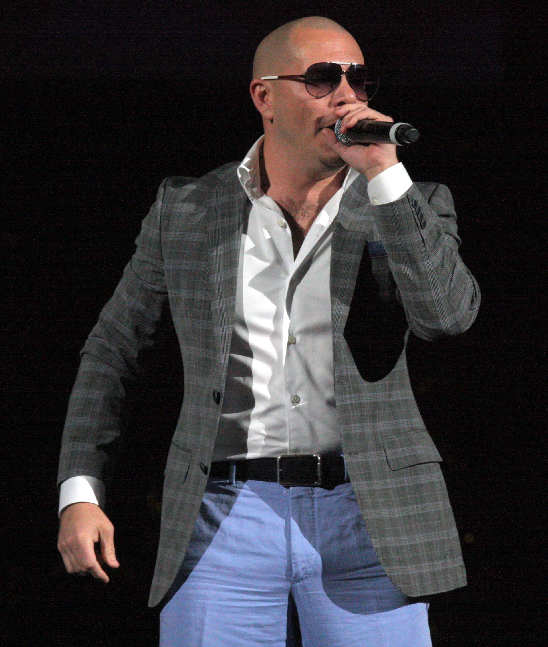 Pitbull, de lentes oscuros y saco a cuadros, canta con el micrófono en la mano sobre un fondo negro.