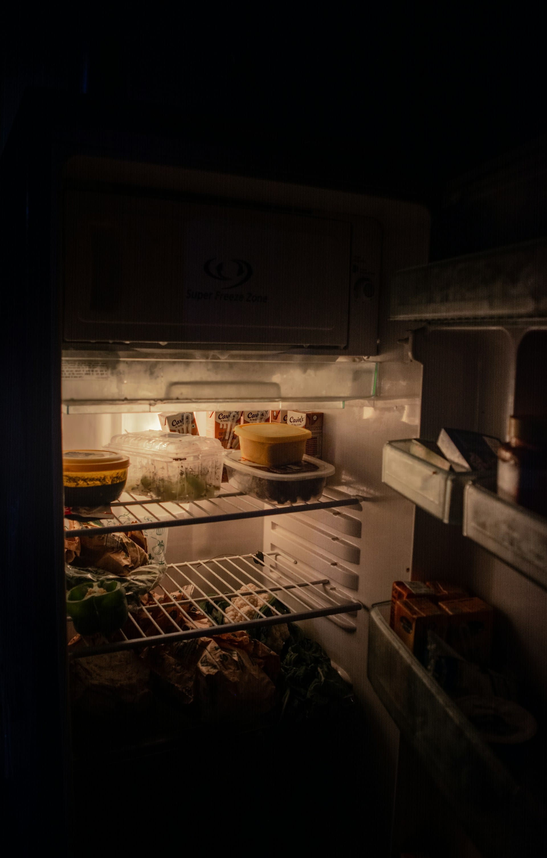 Un refrigerador abierto en una cocina a oscuras, con recipientes y verduras iluminados por su luz interior.