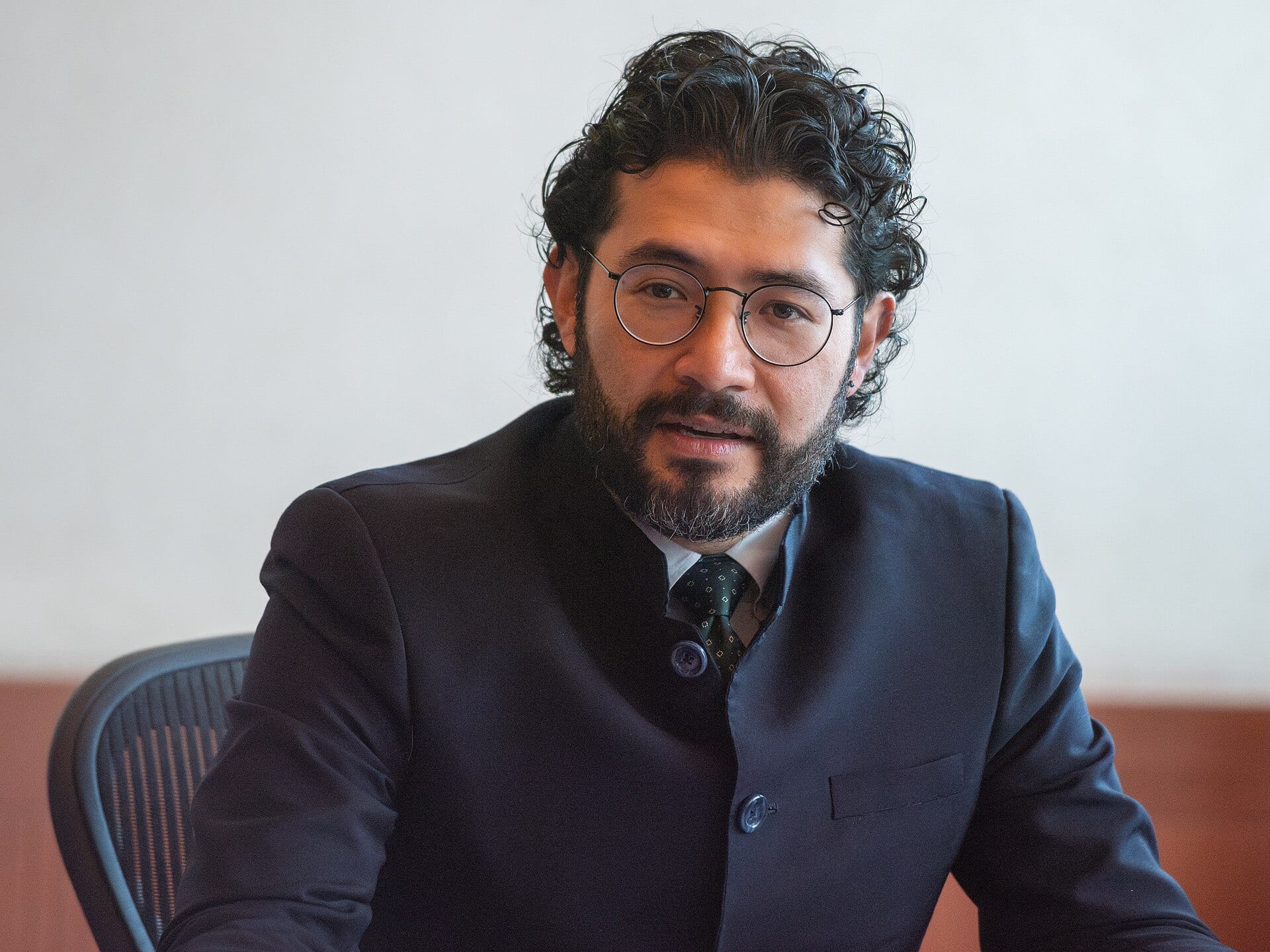 Un hombre de barba, lentes redondos y saco azul oscuro habla sentado frente a una pared clara.