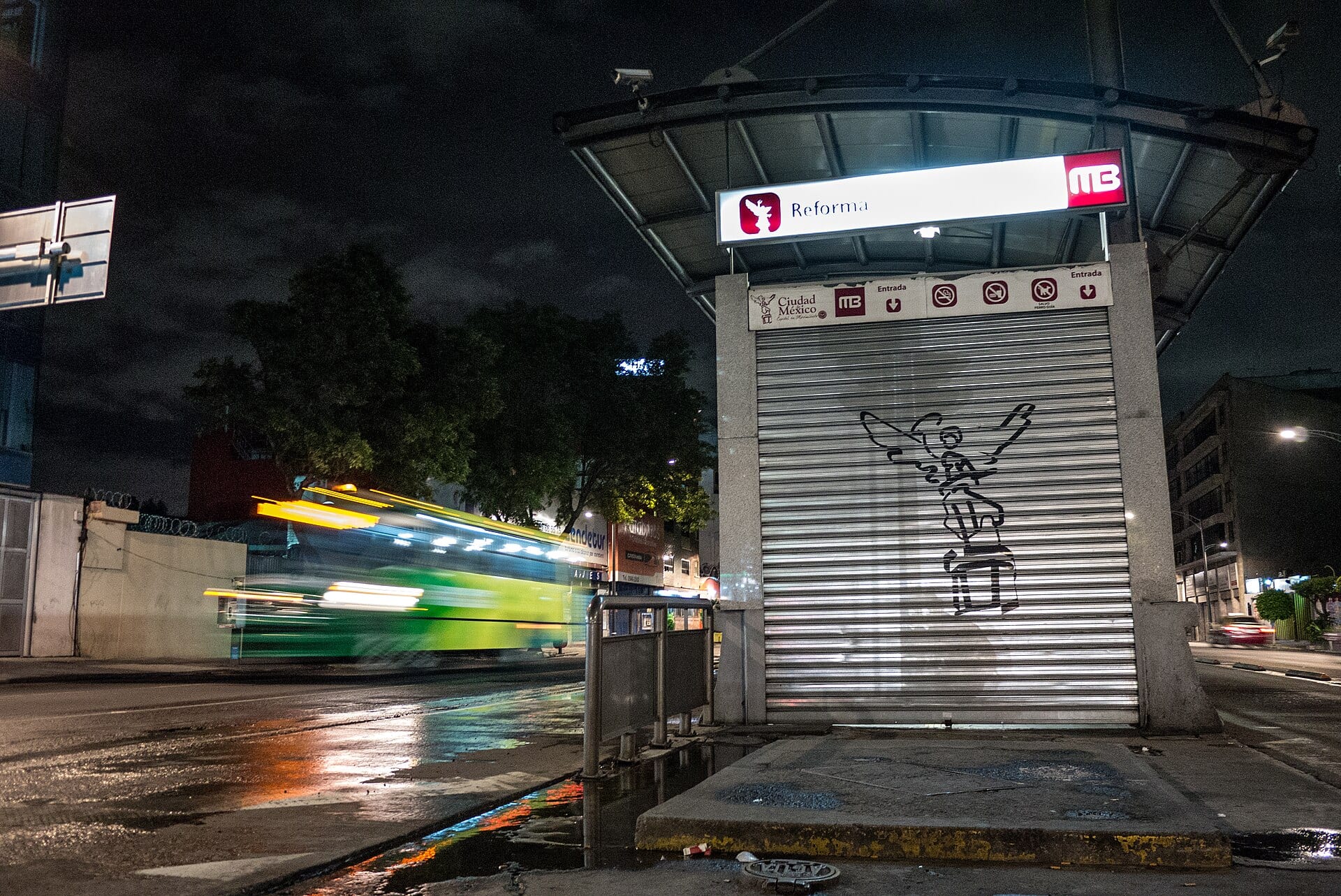 Un autobús verde pasa borroso de noche junto a una estación de Metrobús con el letrero Reforma y la cortina metálica cerrada.