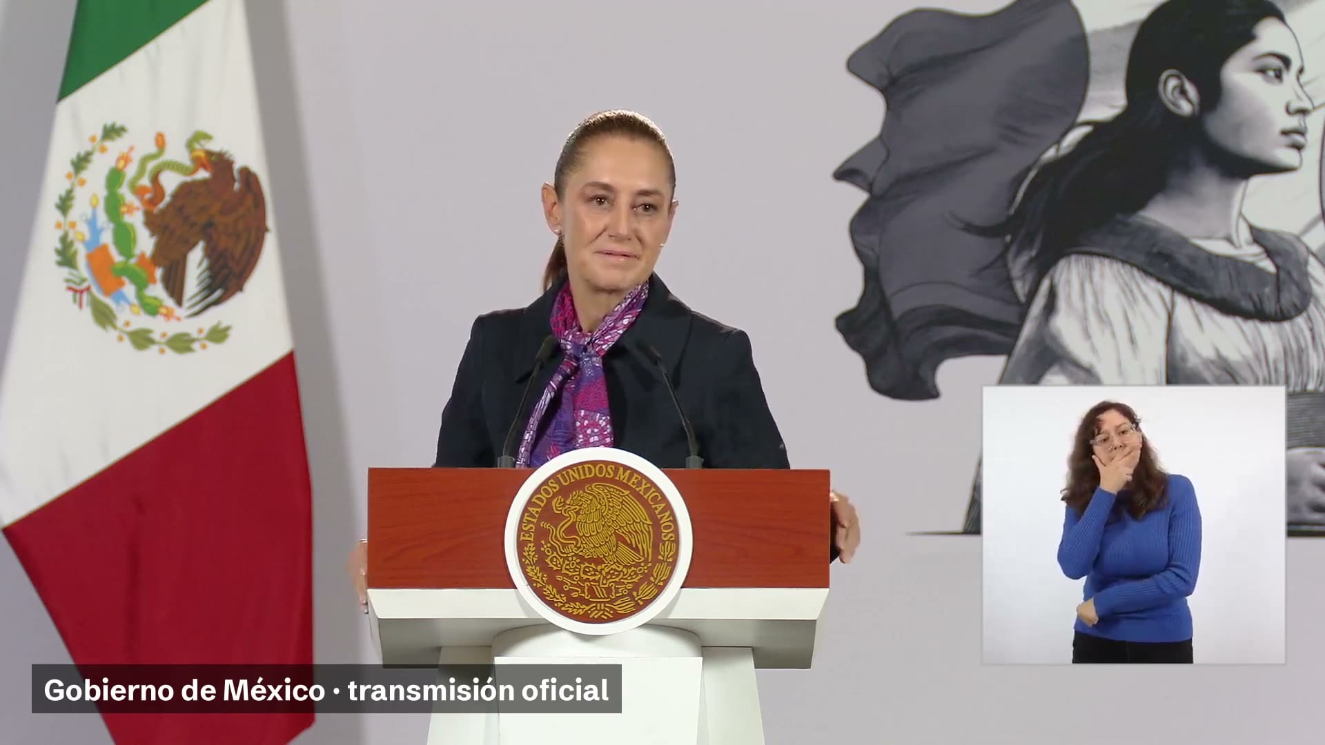 La presidenta Claudia Sheinbaum habla detrás del atril con el escudo nacional, junto a una bandera de México y una intérprete de lengua de señas.