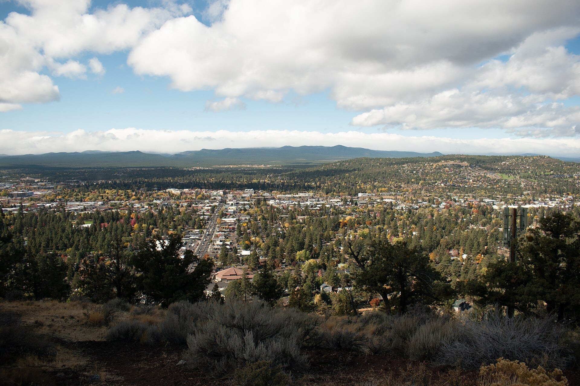 Vista aérea de Bend, Oregón, con una avenida entre casas y pinos y una cadena de montañas al fondo bajo nubes.