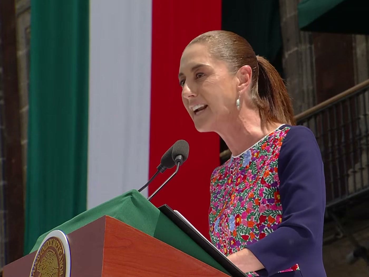 Claudia Sheinbaum habla frente a un micrófono en un atril, con una blusa bordada de flores y una bandera de México detrás.