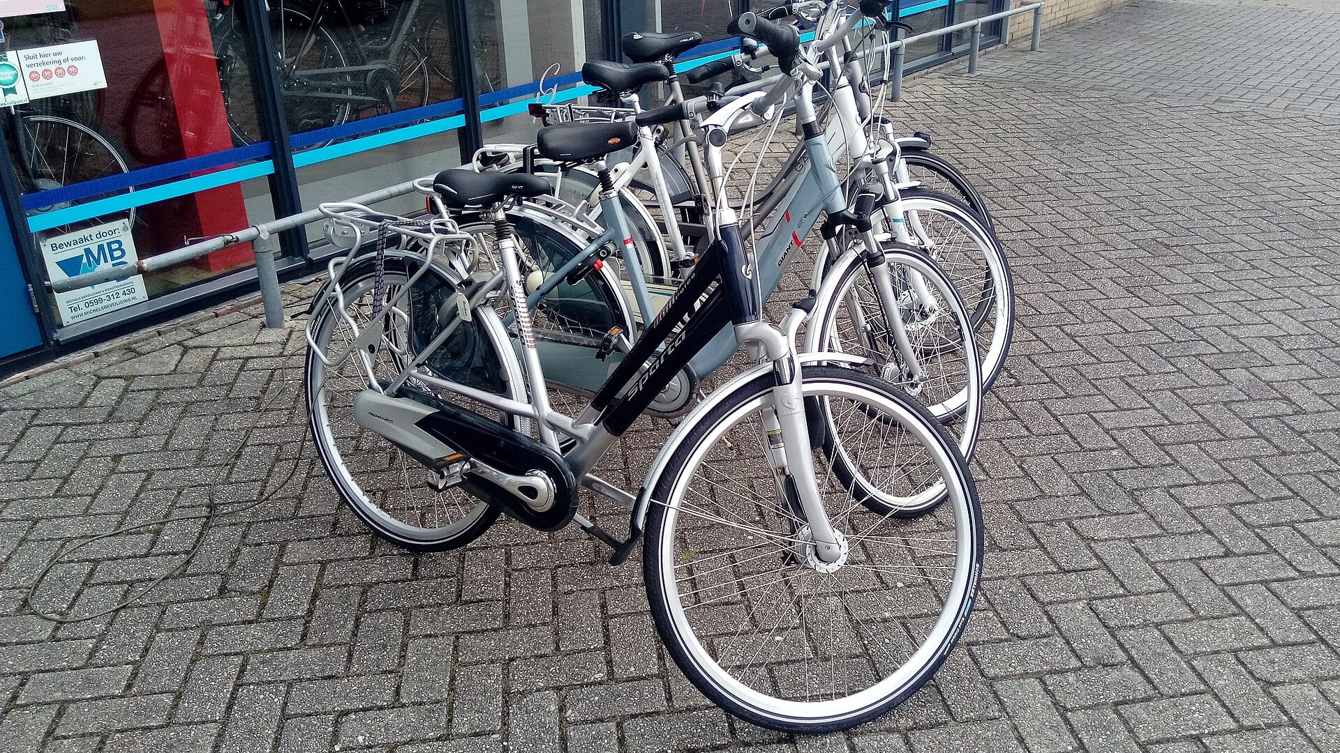 Varias bicicletas de adulto estacionadas en fila frente a un comercio, sobre un piso de adoquín.