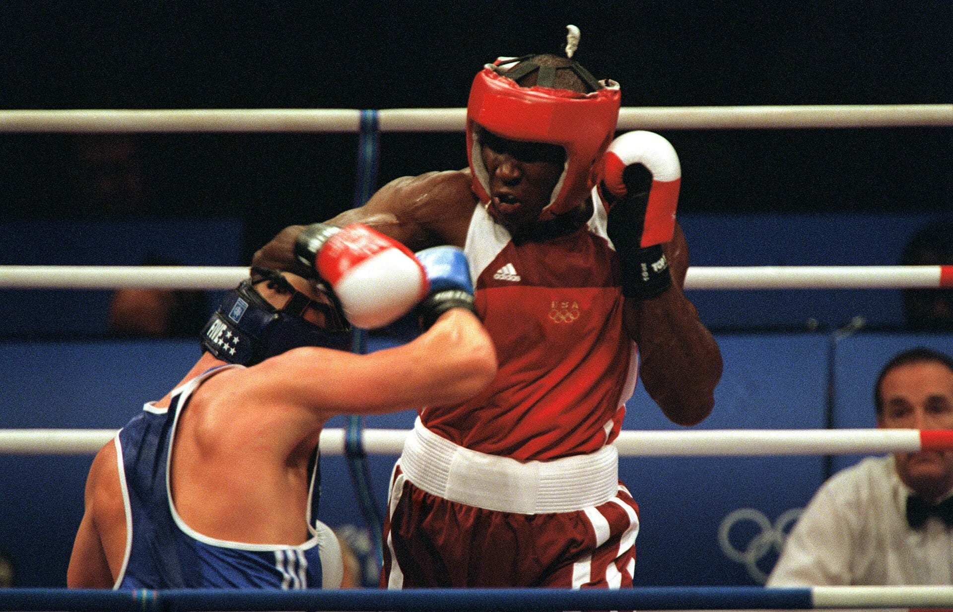 Un boxeador con casco y camiseta roja lanza un golpe a un rival de camiseta azul dentro de un ring.
