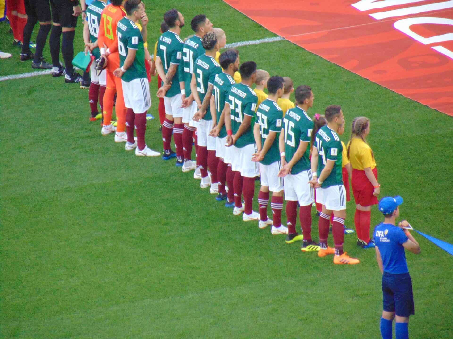 Mexican national team players in green jerseys and white shorts line up with their backs to the camera on the pitch before a match.