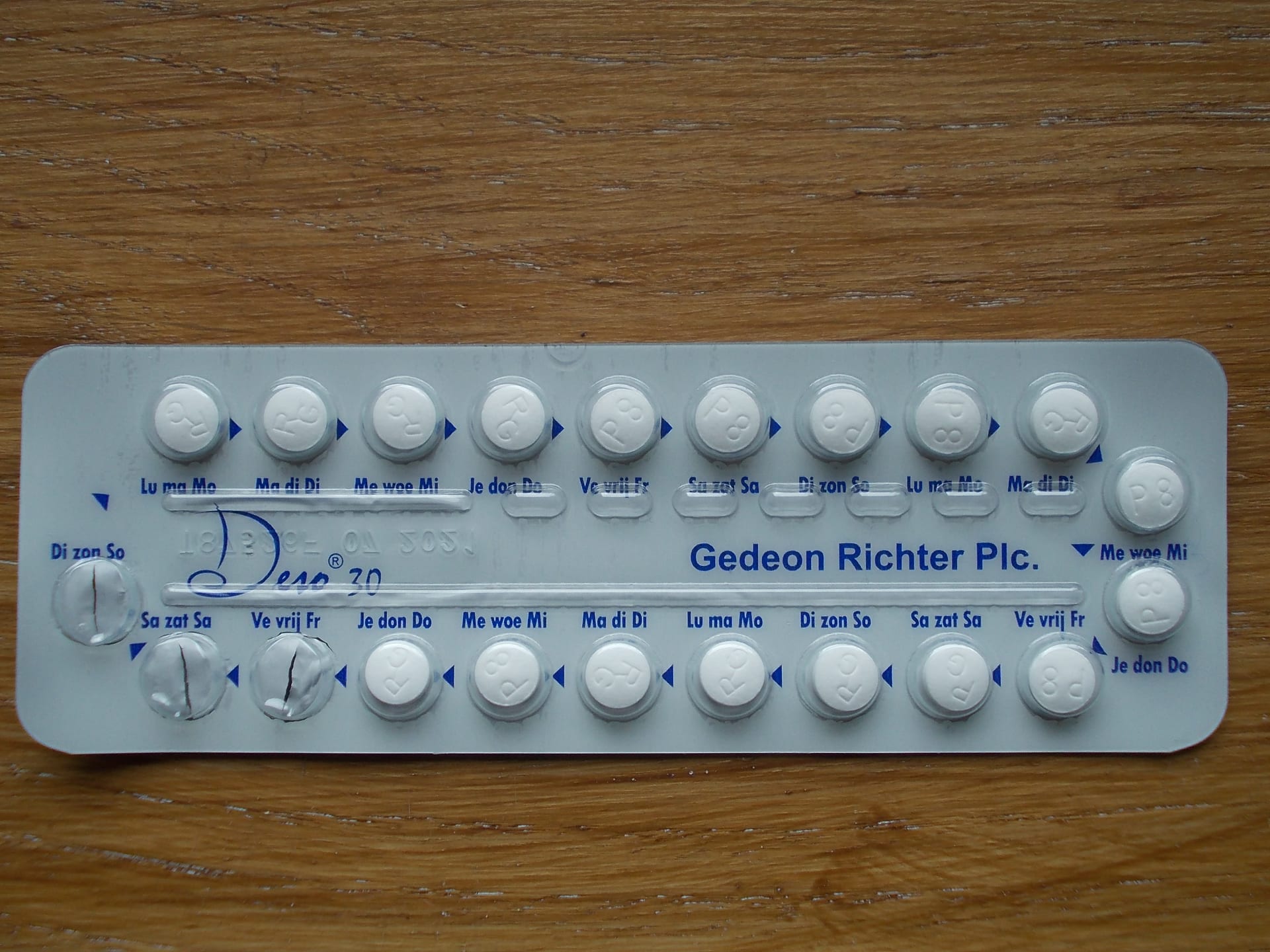 Parte trasera de un blíster plateado de píldoras blancas con días de la semana impresos, sobre una mesa de madera; faltan tres píldoras.