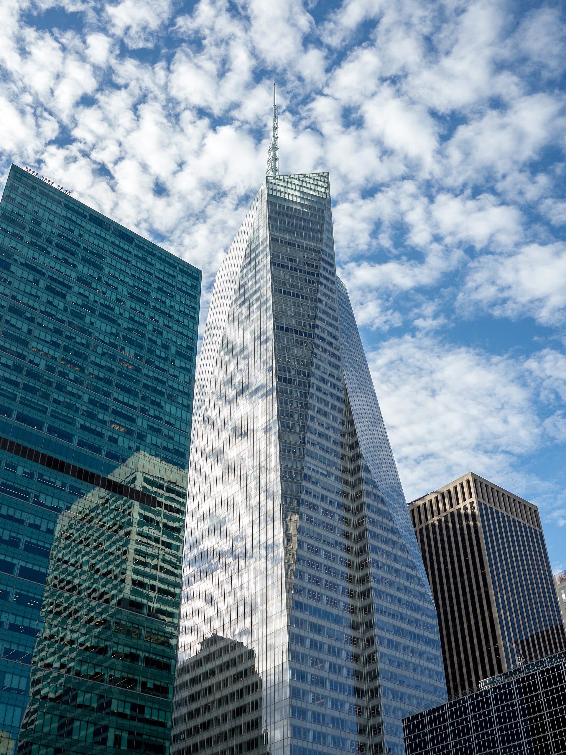 La torre de Bank of America en Manhattan, de fachada de cristal y una antena en la punta, vista desde abajo entre otros edificios y un cielo con nubes.