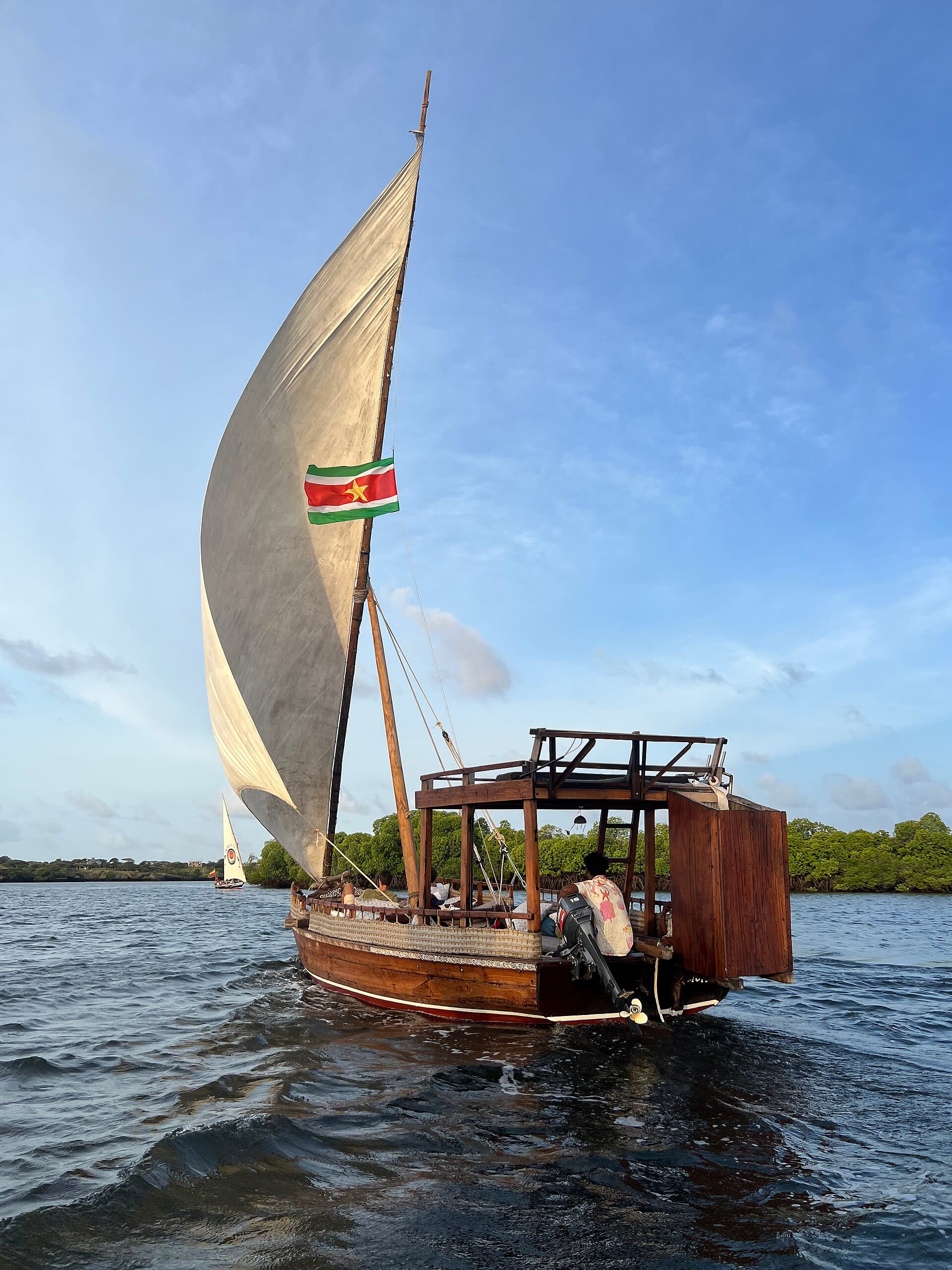 Un velero tradicional de vela triangular navega cerca de la costa, con manglares y un segundo velero al fondo.