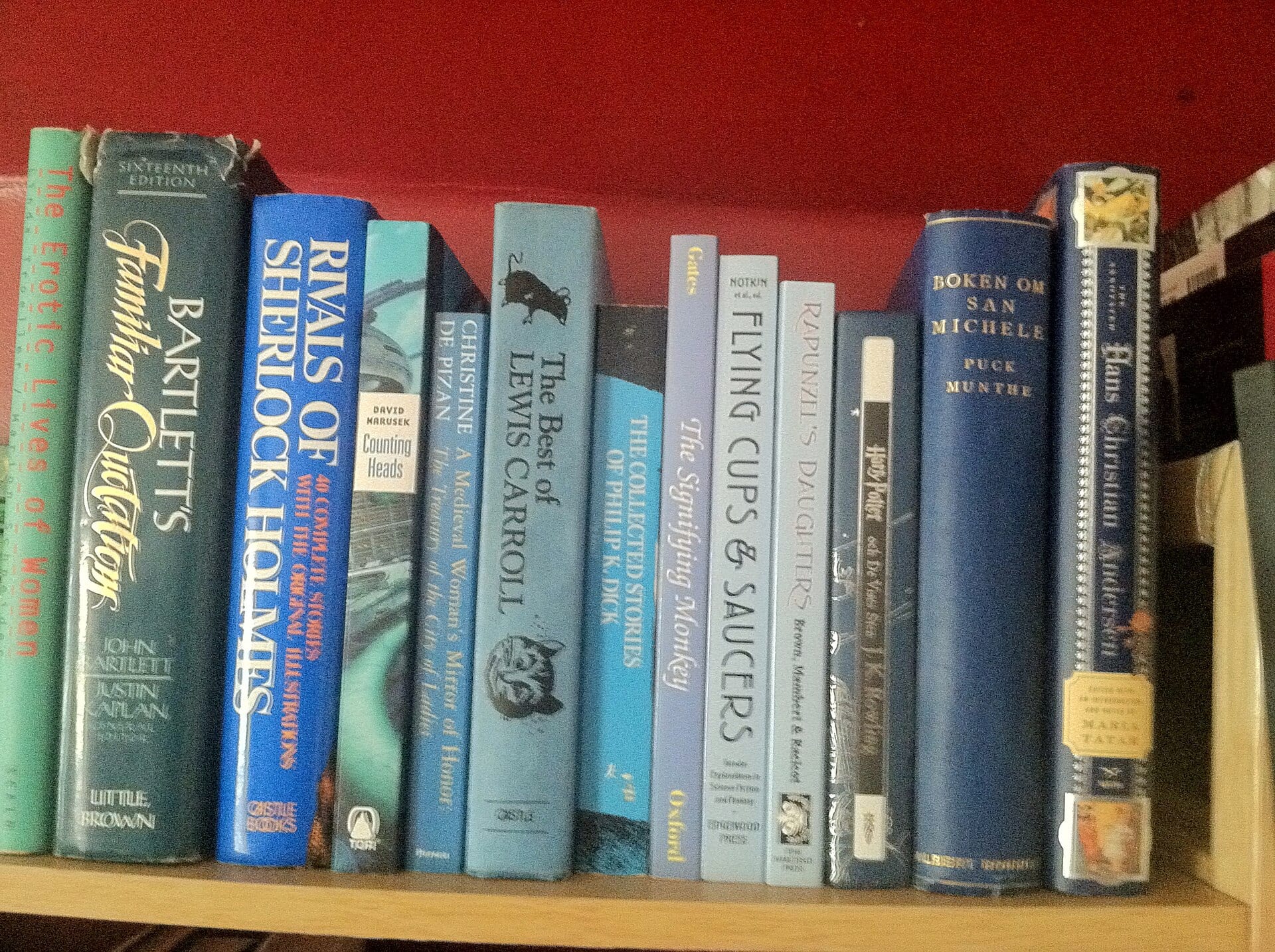 Una hilera de libros de lomo azul y verde sobre un estante de madera, frente a una pared roja.
