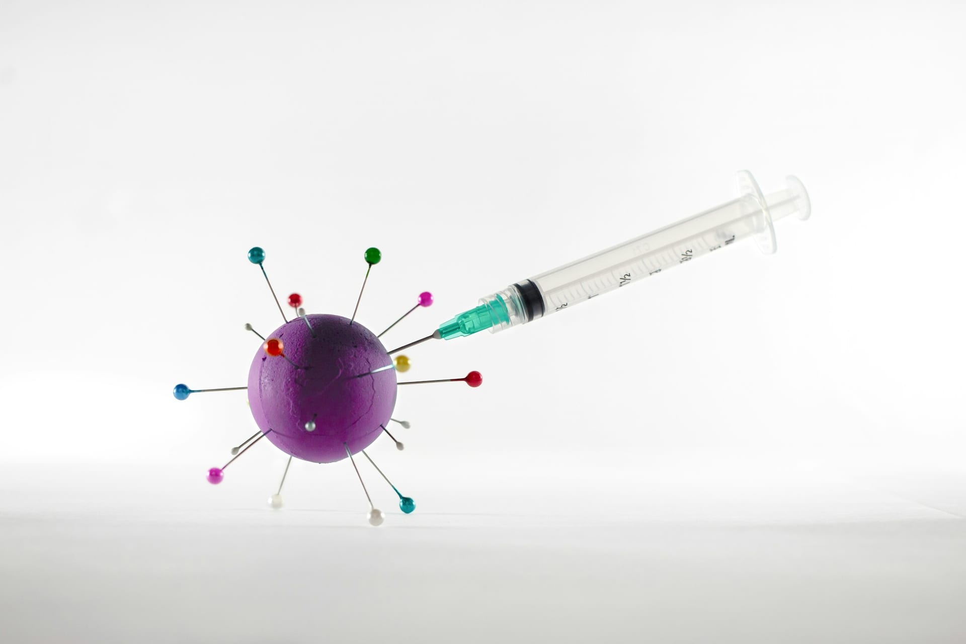 Una jeringa transparente con aguja verde se acerca a una esfera morada con alfileres de colores clavados, sobre fondo blanco.