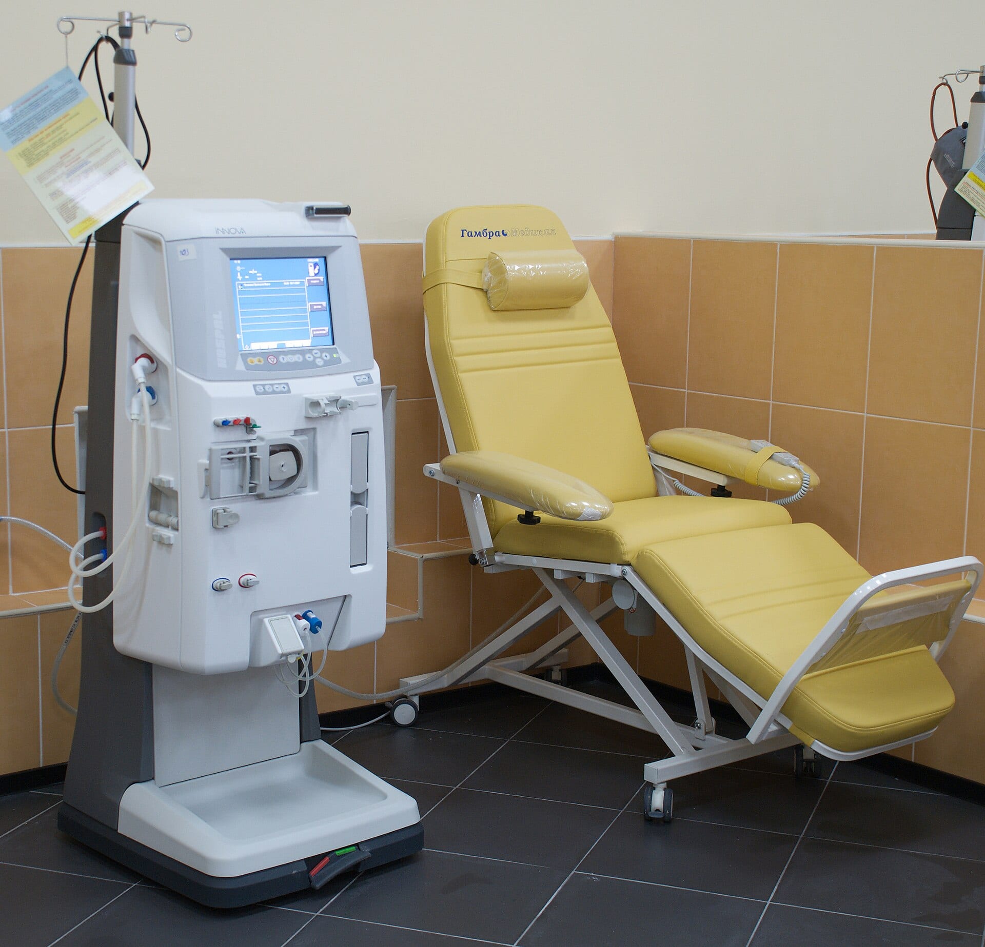 Una máquina de hemodiálisis blanca con pantalla azul junto a un sillón reclinable amarillo en una sala de azulejos color naranja.