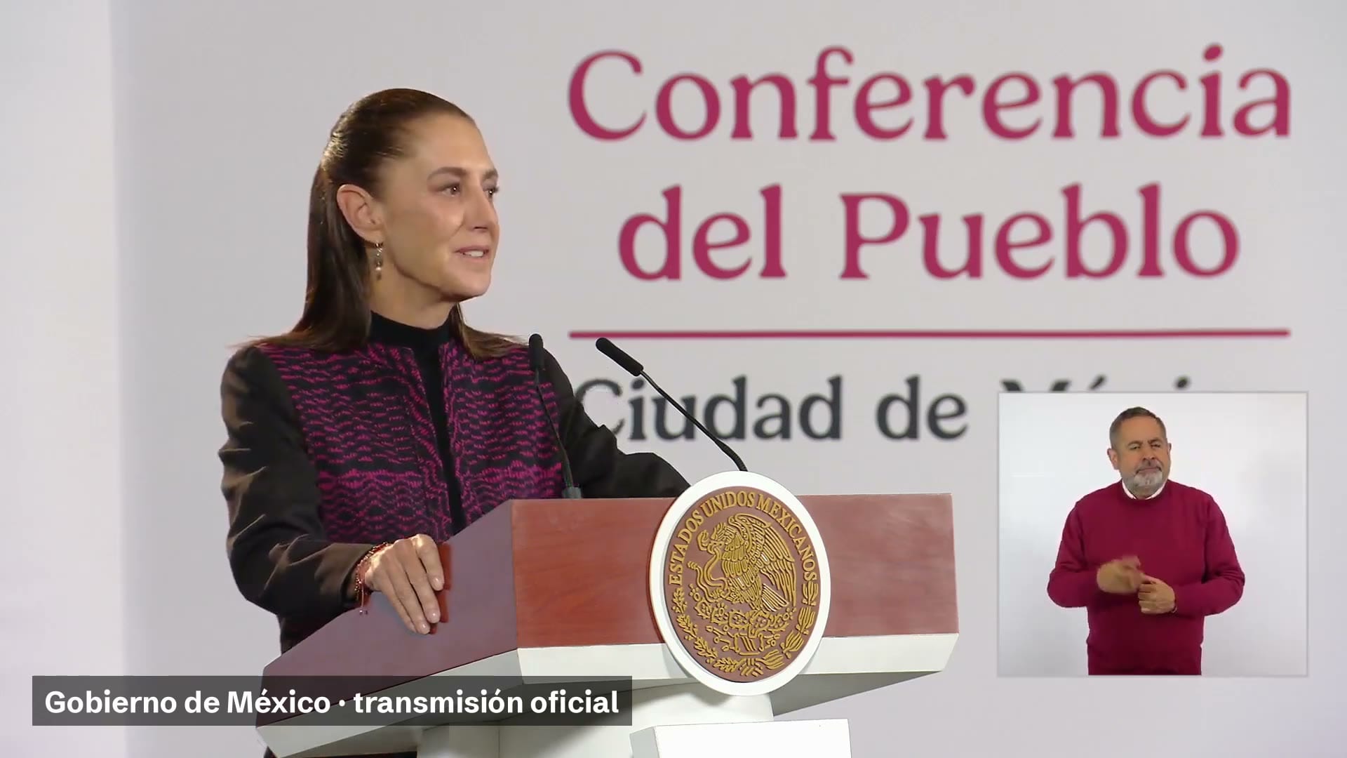 La presidenta Claudia Sheinbaum habla frente al atril con el escudo nacional, junto a una intérprete de lengua de señas.