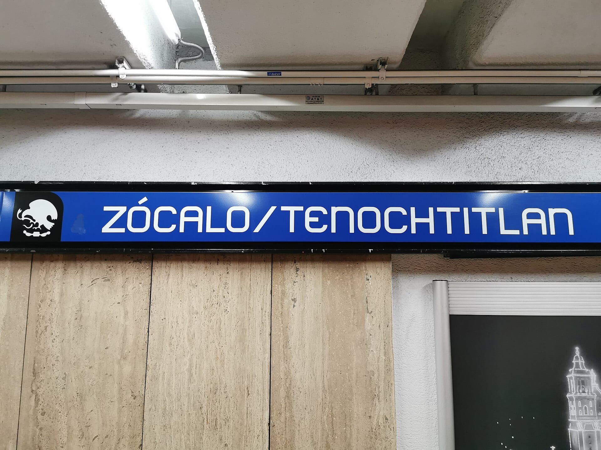 El letrero azul de la estación Zócalo/Tenochtitlan del Metro, con su logo, sobre un pasillo de muros de piedra.