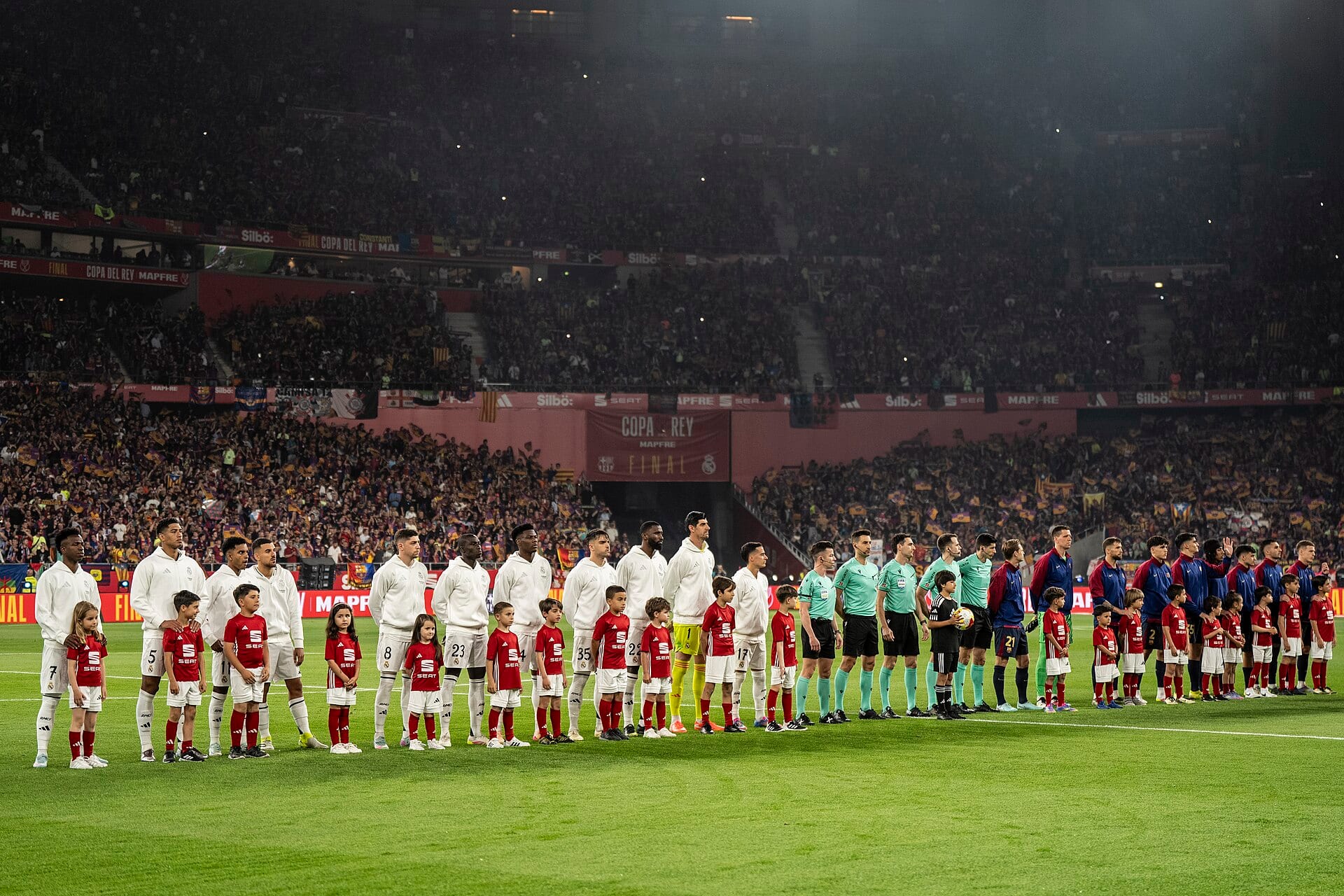Real Madrid players in white warm-up tops lined up beside children in red shirts and the four match officials before a Copa del Rey final.