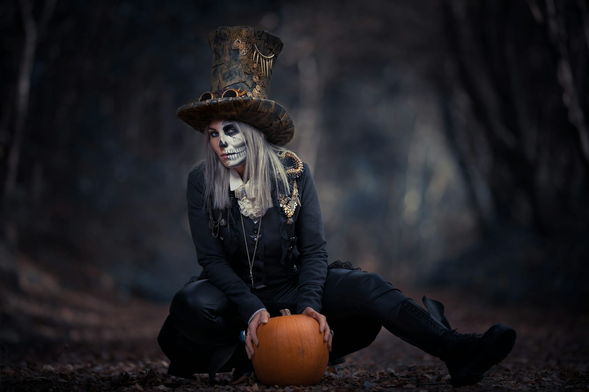 Una mujer con maquillaje de esqueleto en la cara y un sombrero de copa de estilo steampunk sostiene una calabaza sobre un camino de hojas secas.