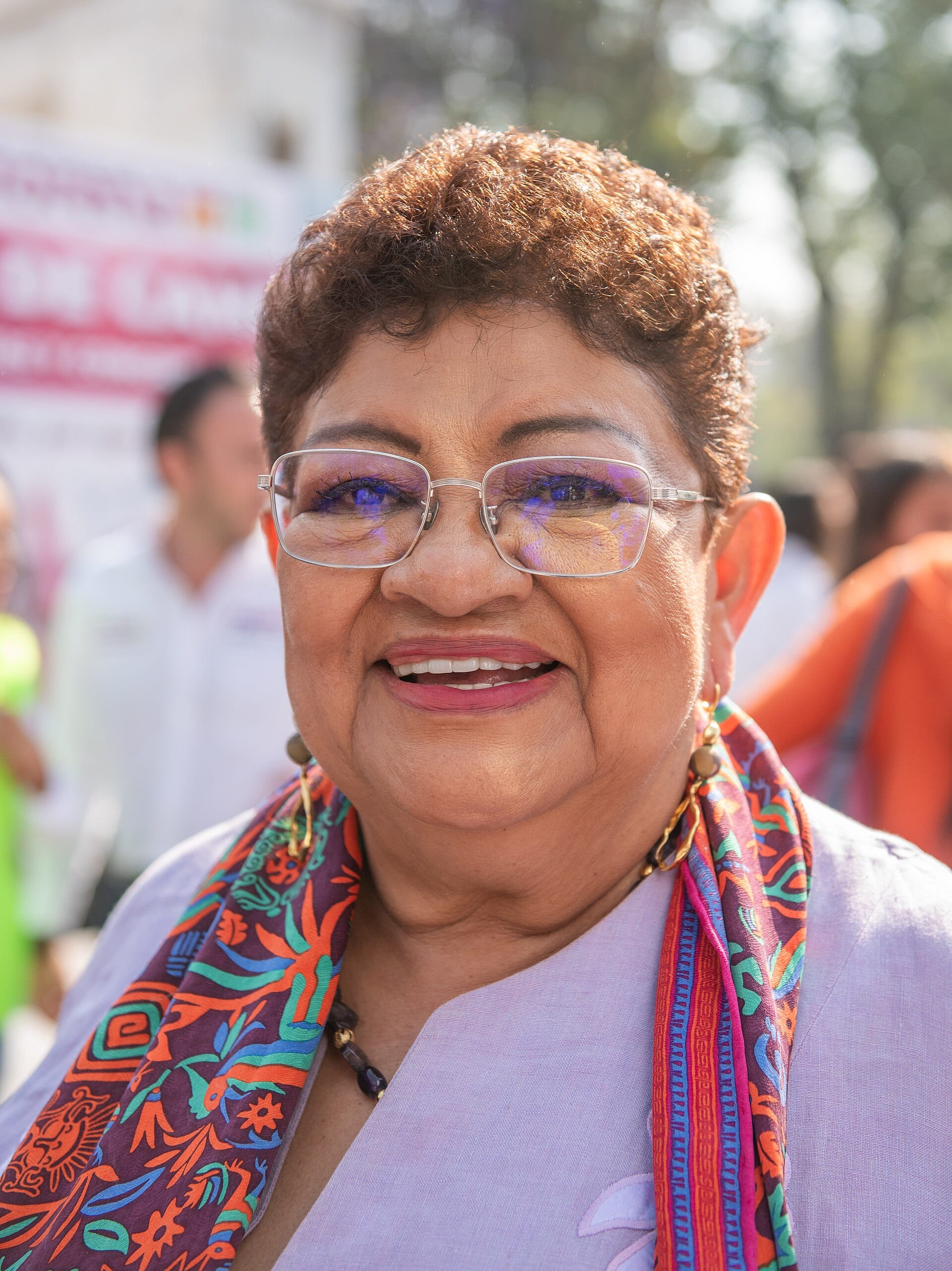 Primer plano de Ernestina Godoy Ramos sonriendo, con lentes, cabello corto y un pañuelo estampado sobre una blusa lila, al aire libre.