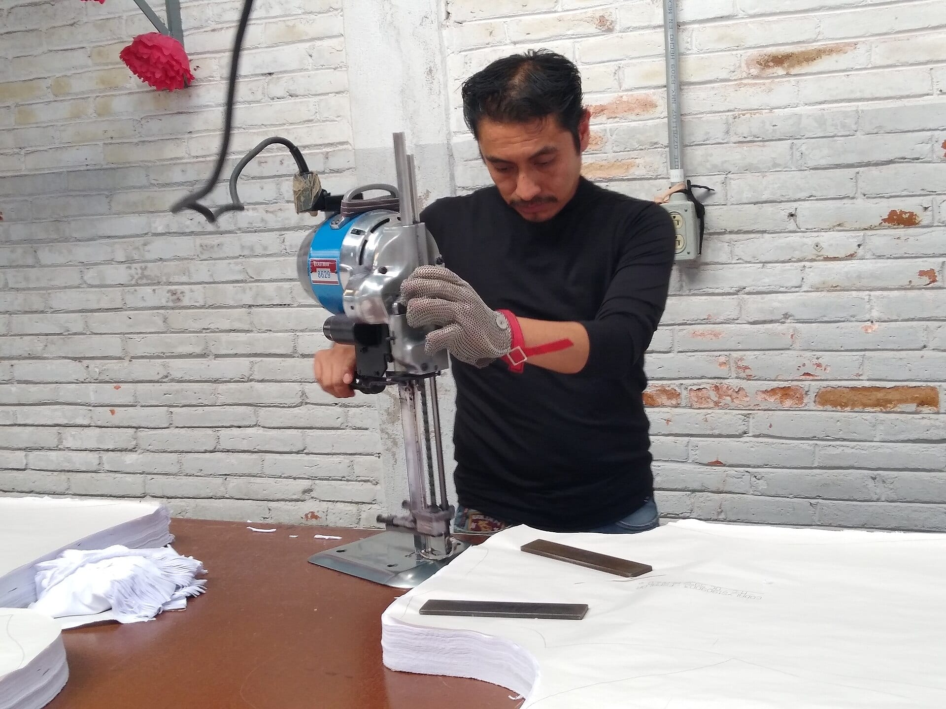 Un operario con guante de malla metálica guía una cortadora vertical sobre capas de tela blanca apiladas en una mesa.