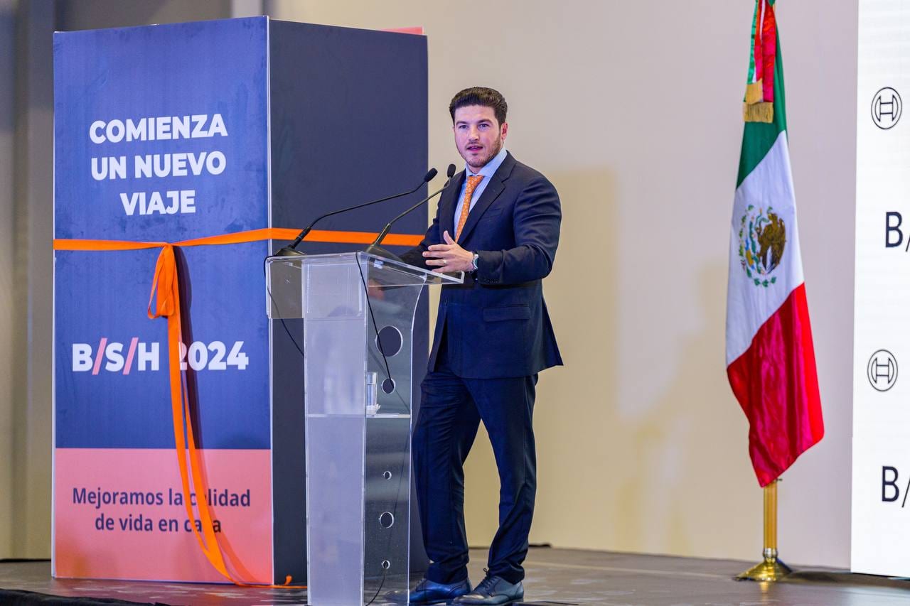 Samuel García habla desde un podio de acrílico, con una bandera de México a su derecha y un cartel azul con una cinta naranja a su izquierda.