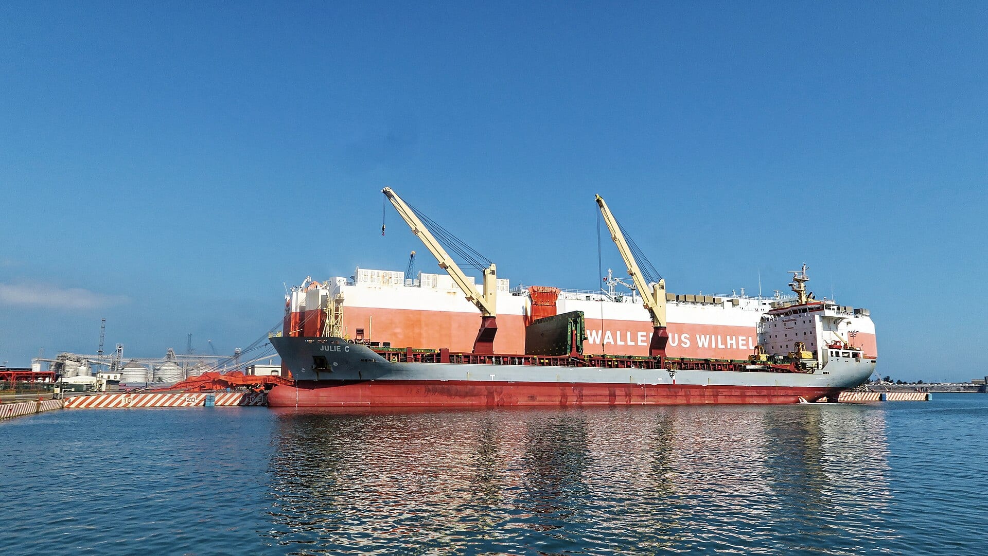 Un buque de carga con dos grúas amarrado a un muelle de franjas rojas y blancas, bajo un cielo azul, en el puerto de Veracruz.