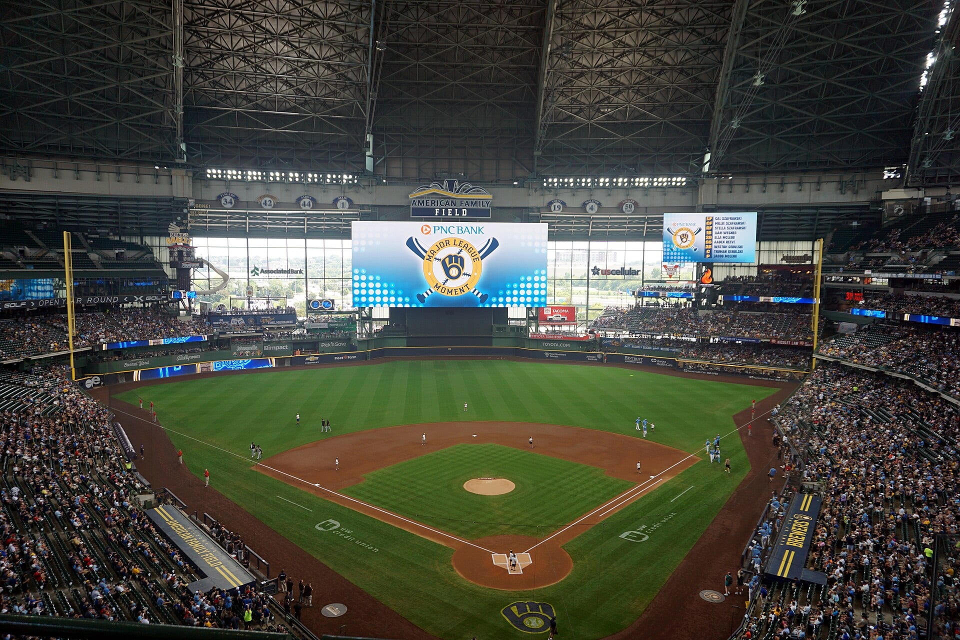 Vista del diamante del American Family Field desde las gradas altas, con el techo abierto, la pantalla central y público en los asientos.