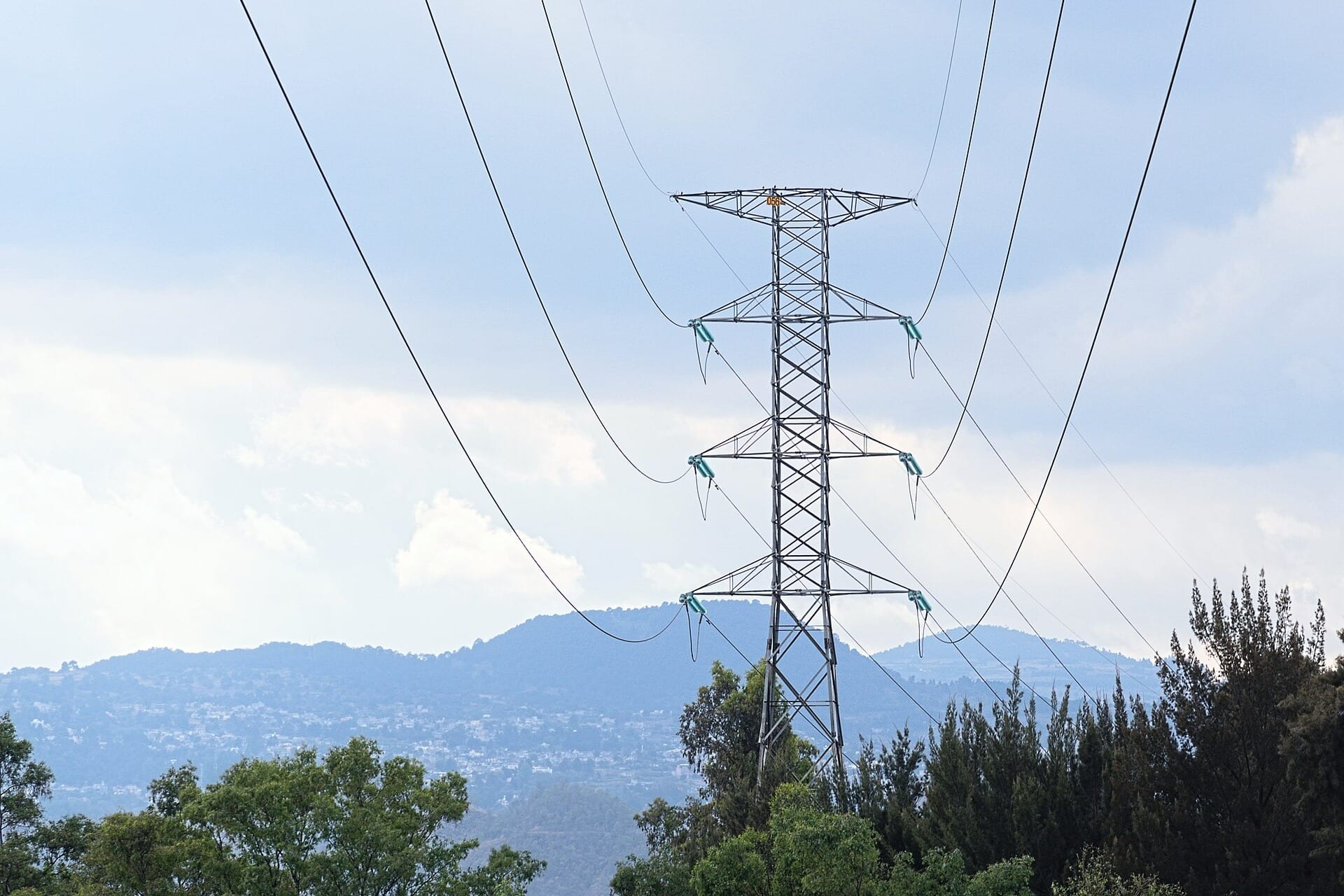 Una torre de transmisión eléctrica con sus cables entre árboles, frente a un cuerpo de agua y unos cerros al fondo.