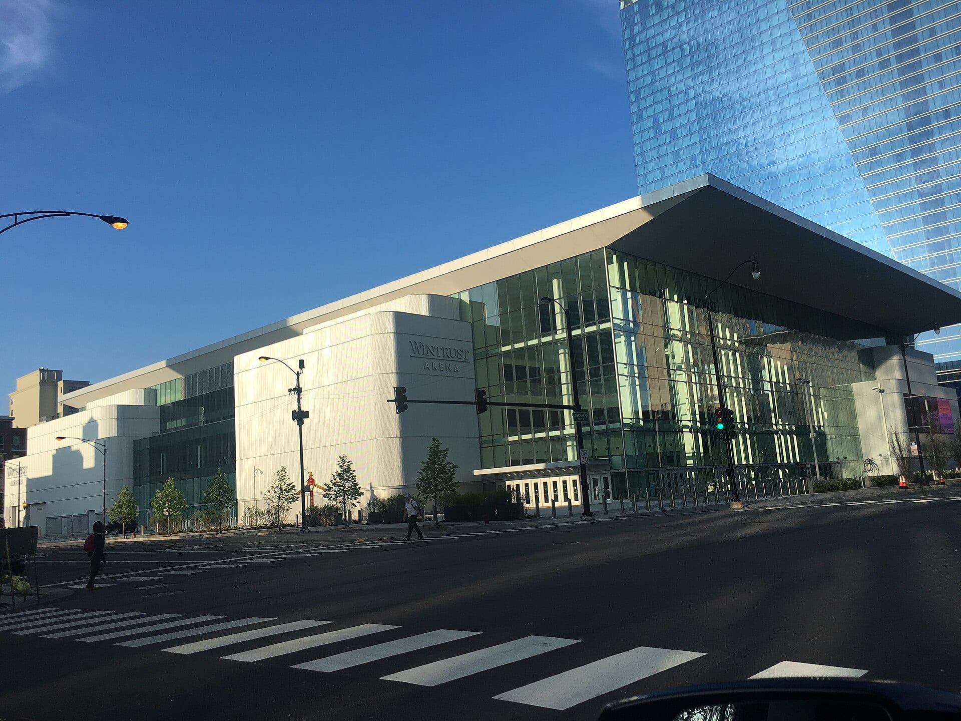 The glass and white-paneled facade of Wintrust Arena with the venue's sign, seen from a street corner under a blue sky.