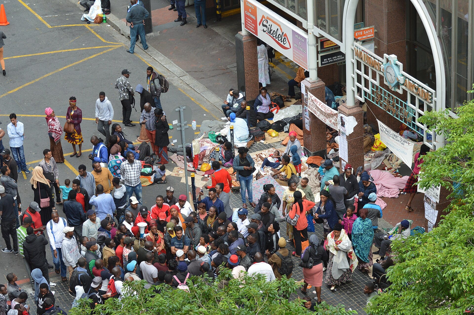 Una multitud se reúne en una calle de Ciudad del Cabo junto a mantas y pertenencias en el suelo, con una manta que pide dejar Sudáfrica colgada de una reja.