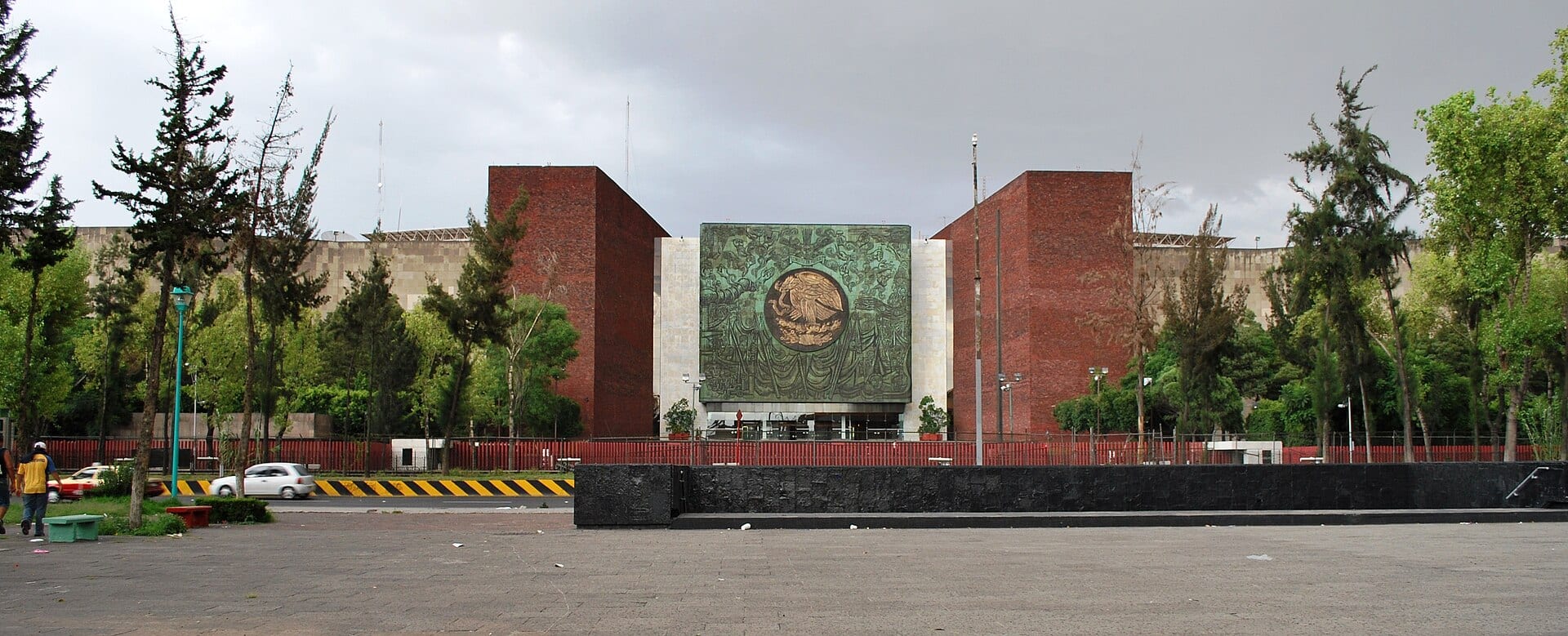 Fachada del Palacio Legislativo de San Lázaro, con un mural verde del escudo nacional entre dos torres de ladrillo rojo y árboles al frente.