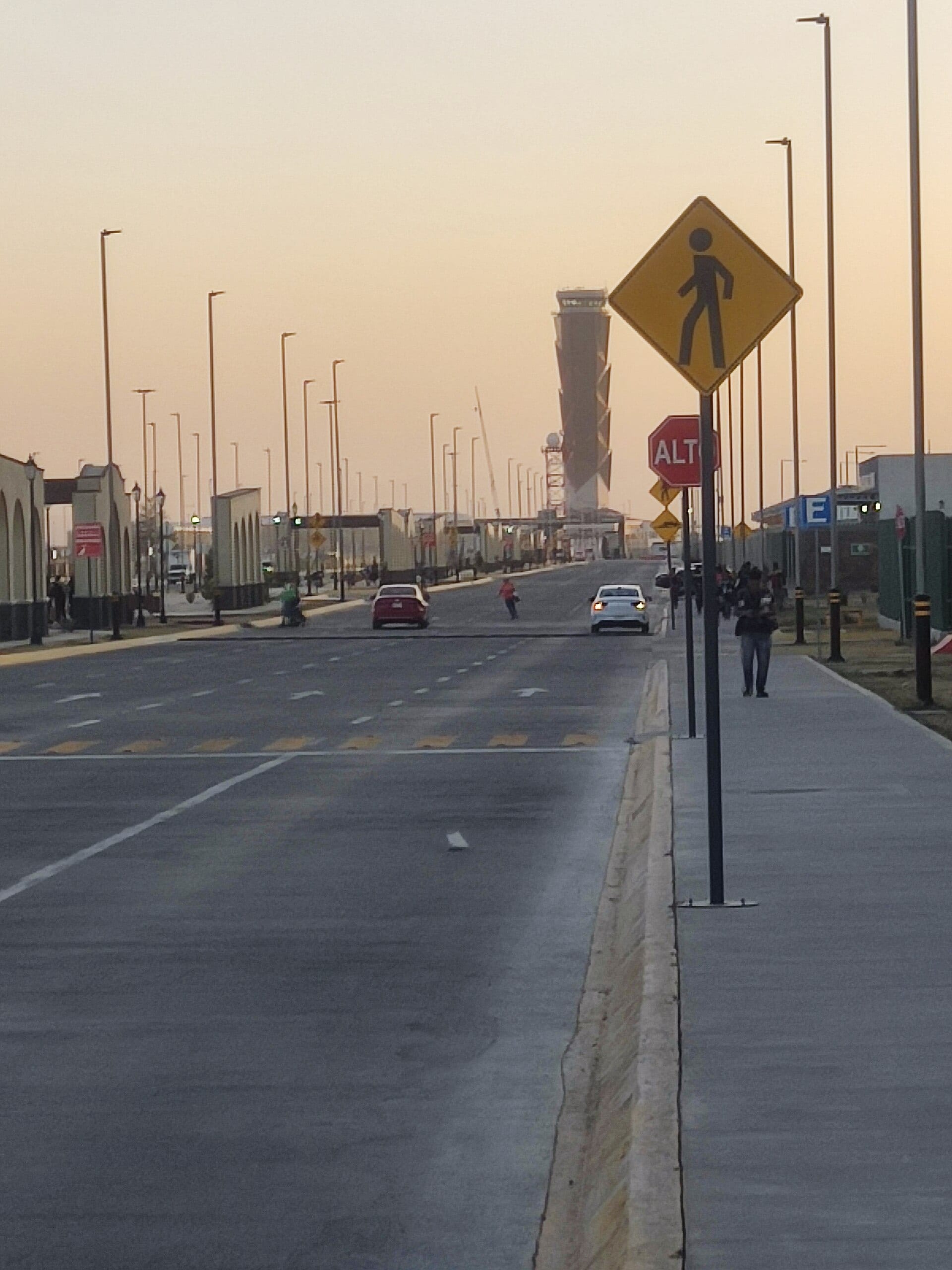 Una avenida con luminarias y señales de tránsito conduce hacia la torre de control del aeropuerto al atardecer.