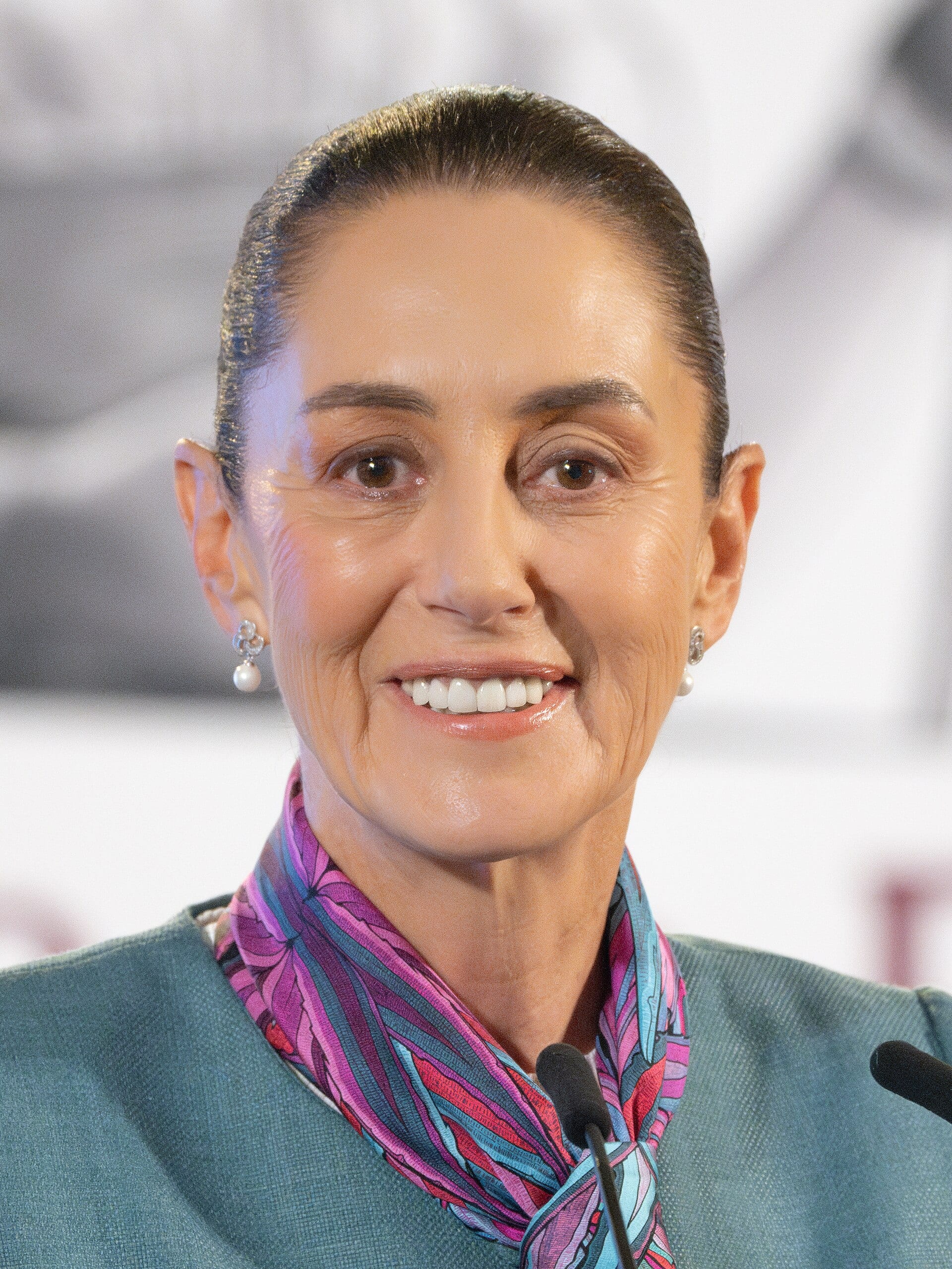 La presidenta Claudia Sheinbaum, de frente y sonriendo, habla ante un micrófono con un pañuelo estampado al cuello.
