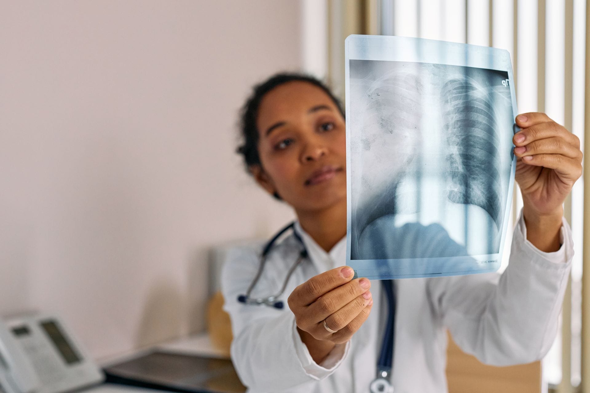 Una médica con bata blanca y estetoscopio sostiene a contraluz una radiografía de tórax en un consultorio.