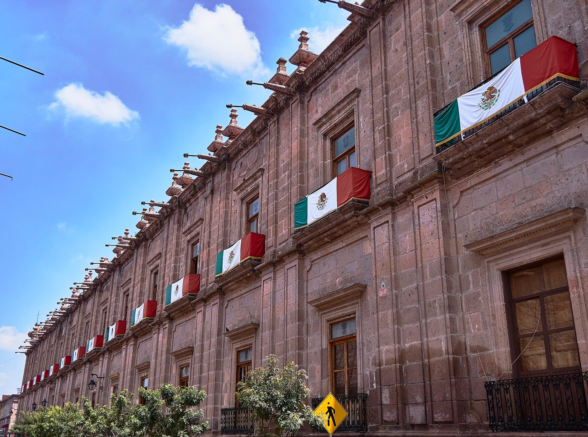 Fachada de cantera del Palacio de Gobierno de Morelia, con banderas de México colgadas de los balcones bajo un cielo azul.