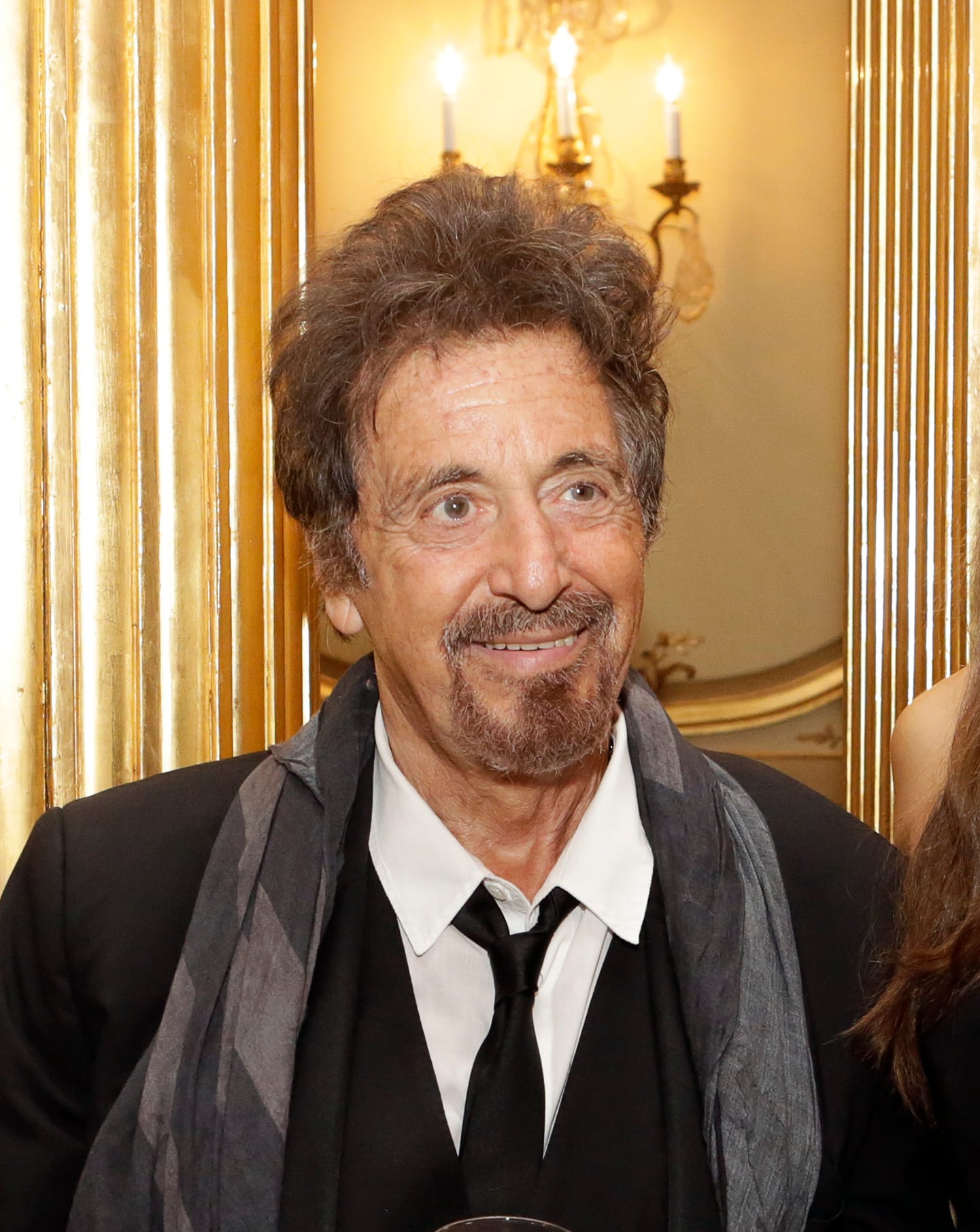 Al Pacino sonríe con chivo y cabello revuelto, con saco negro, corbata y bufanda gris, frente a una pared dorada con candelabros encendidos.
