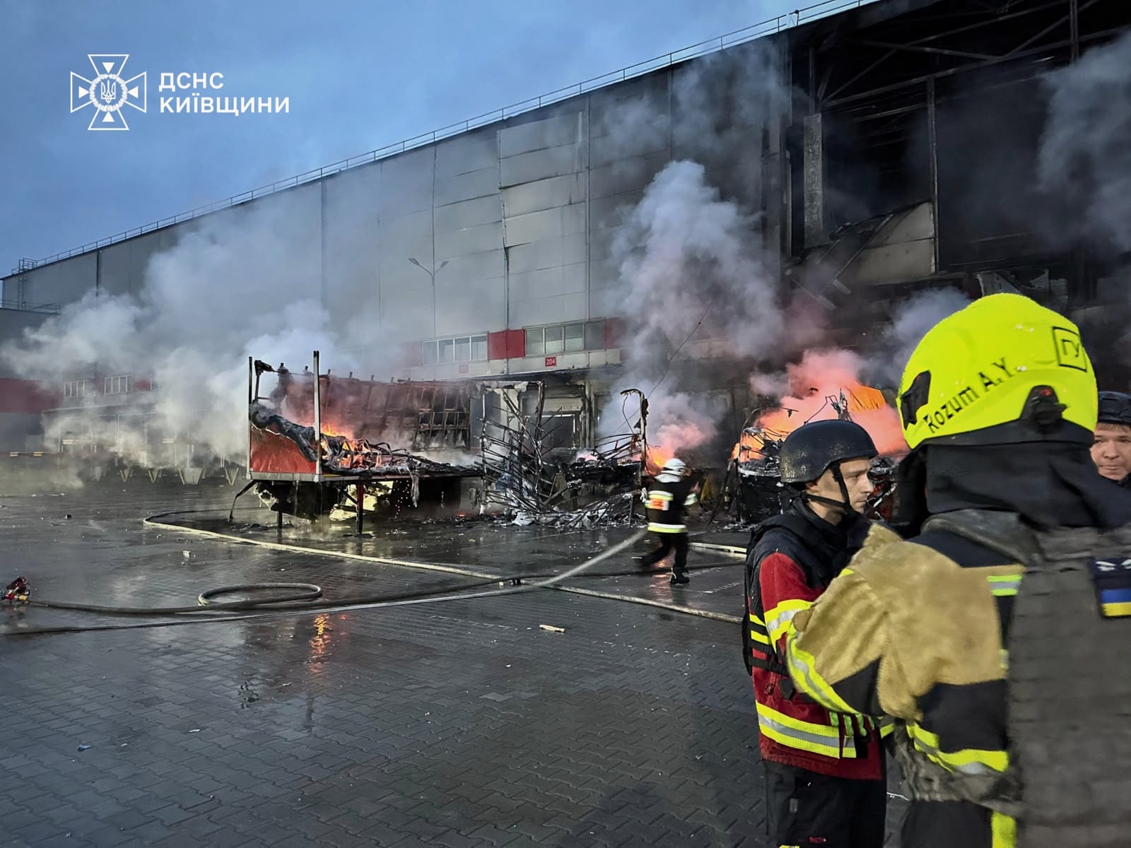 Bomberos con casco amarillo frente a una bodega humeante y un remolque en llamas.