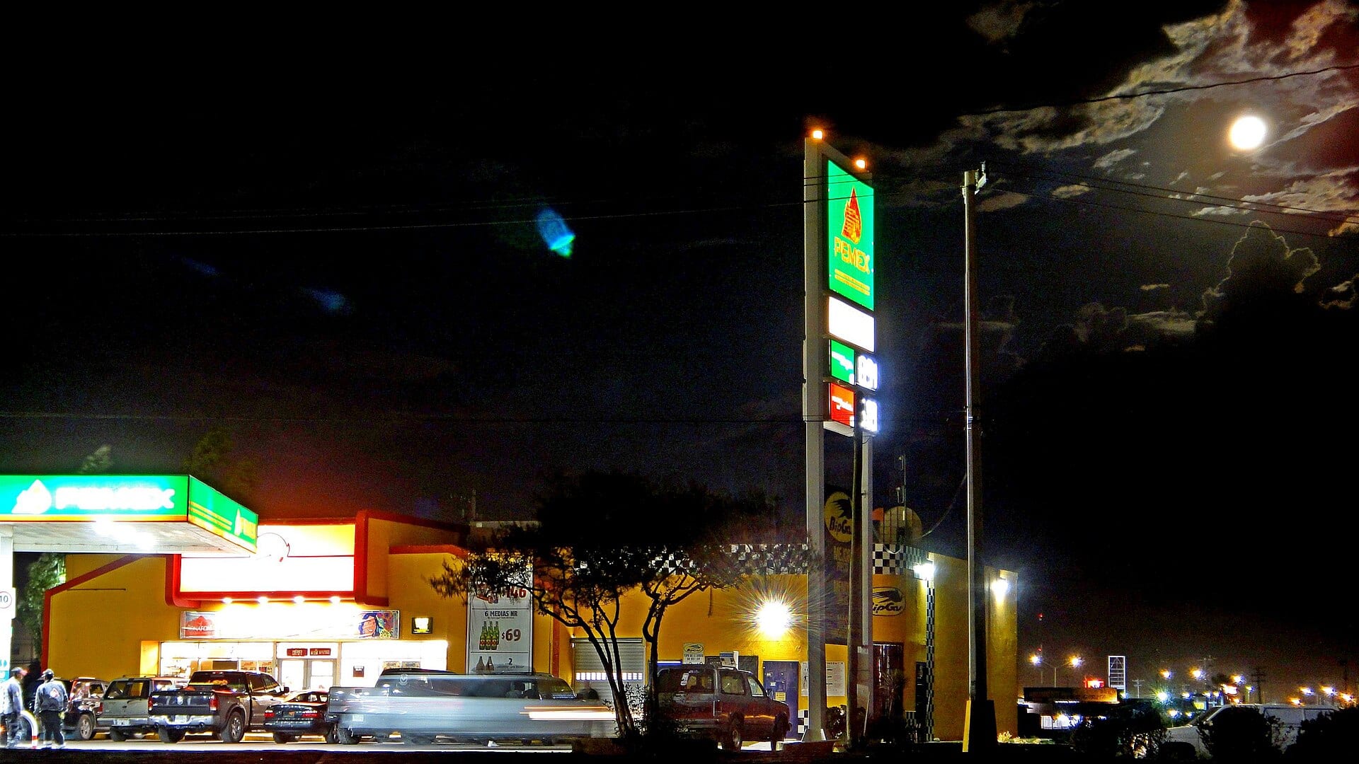Gasolinera Pemex de noche, con el anuncio de precios iluminado, camionetas junto a la tienda y la luna llena entre las nubes.
